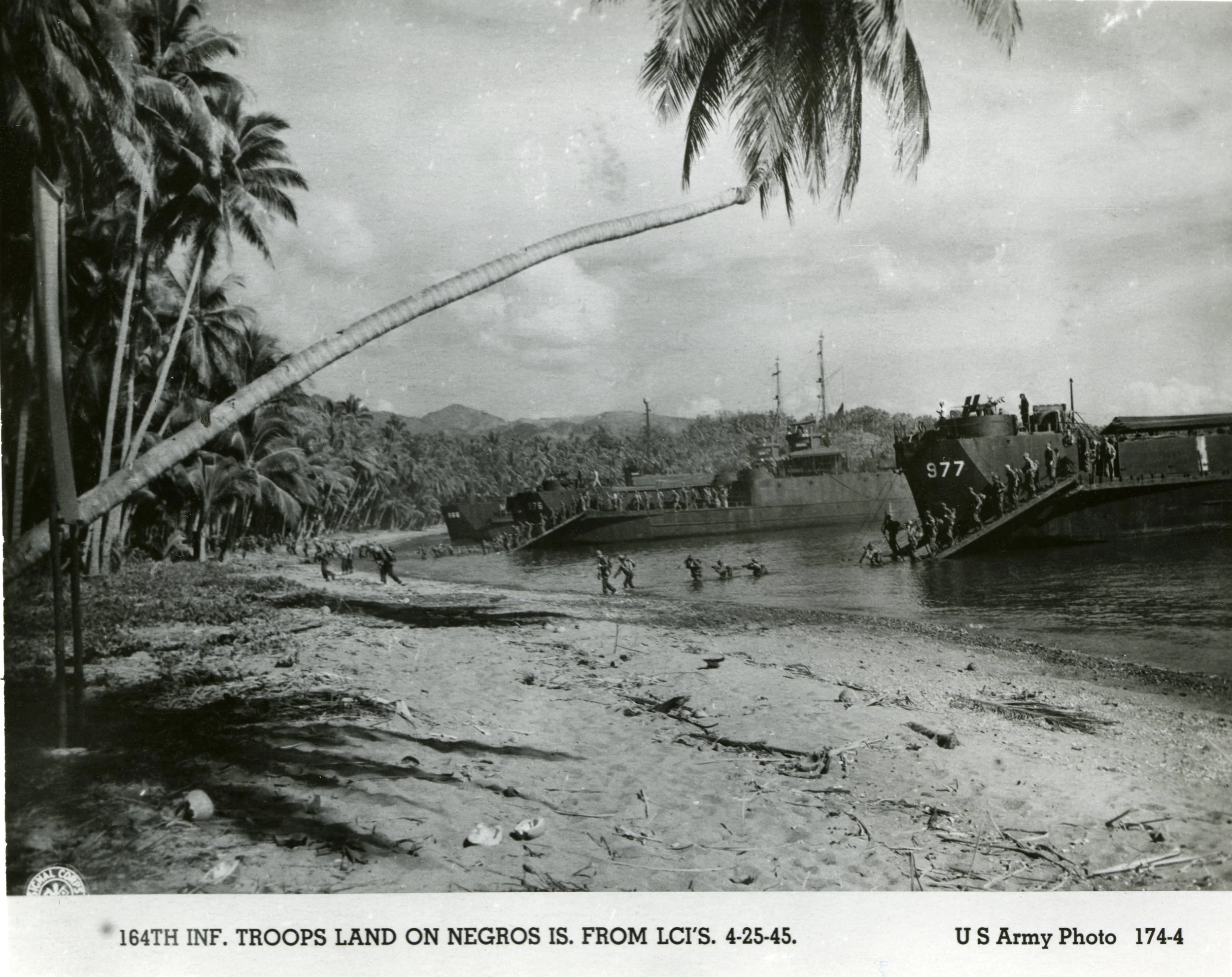 Black and white photo of military troops disembarking from ships onto a tropical beach with palm trees, with mountains in the background. The soldiers are wading through water and climbing onto the shore.