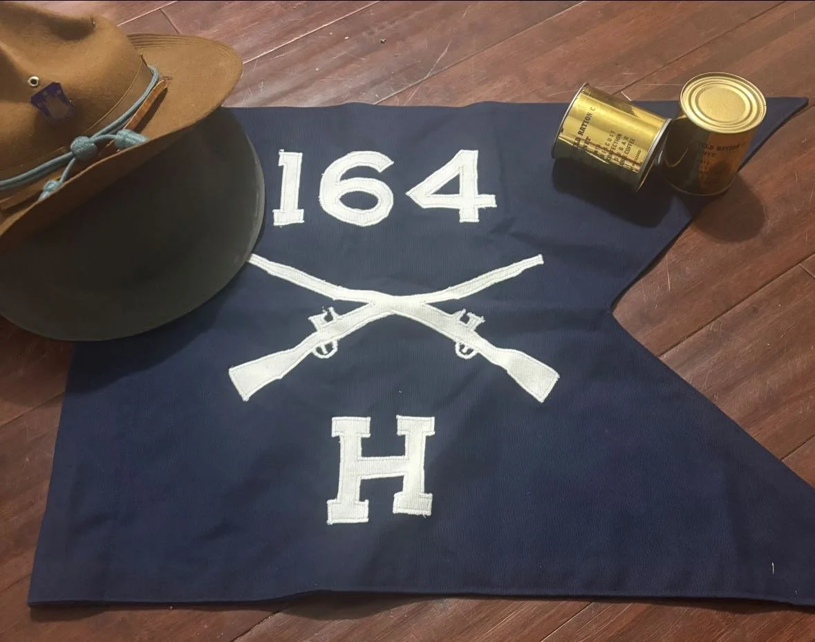 A  blue Infantry with white embroidery featuring the number 164, crossed rifles, and the letter H, placed on a wooden floor. To the left, there is a brown campaign hat. To the right, there are two C-Ration cans stacked on their sides.