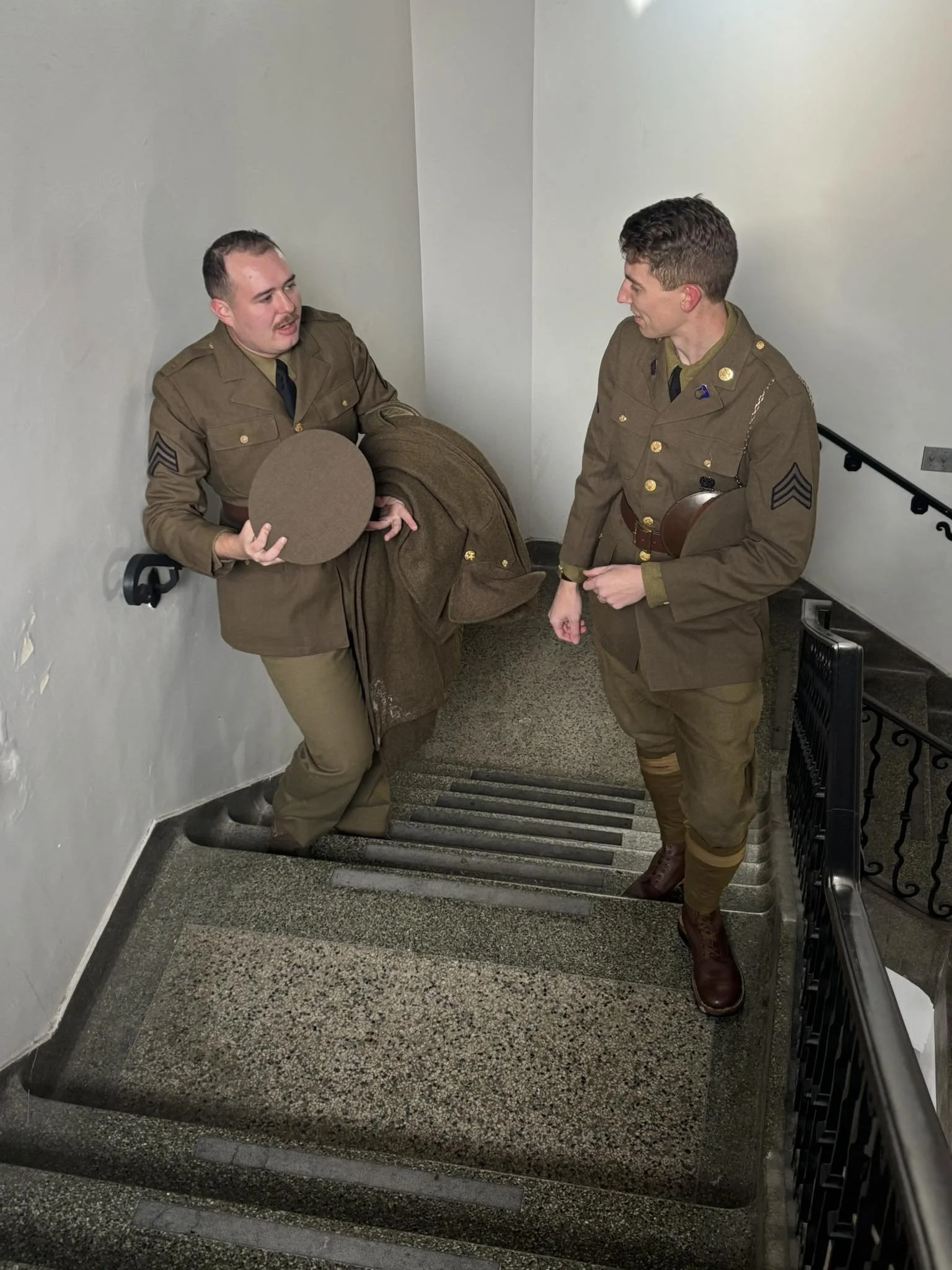 Two living historians in military uniforms engaged in conversation on a staircase, one holding a hat and a coat, the other with a belt and insignia on his uniform, early February 1941.