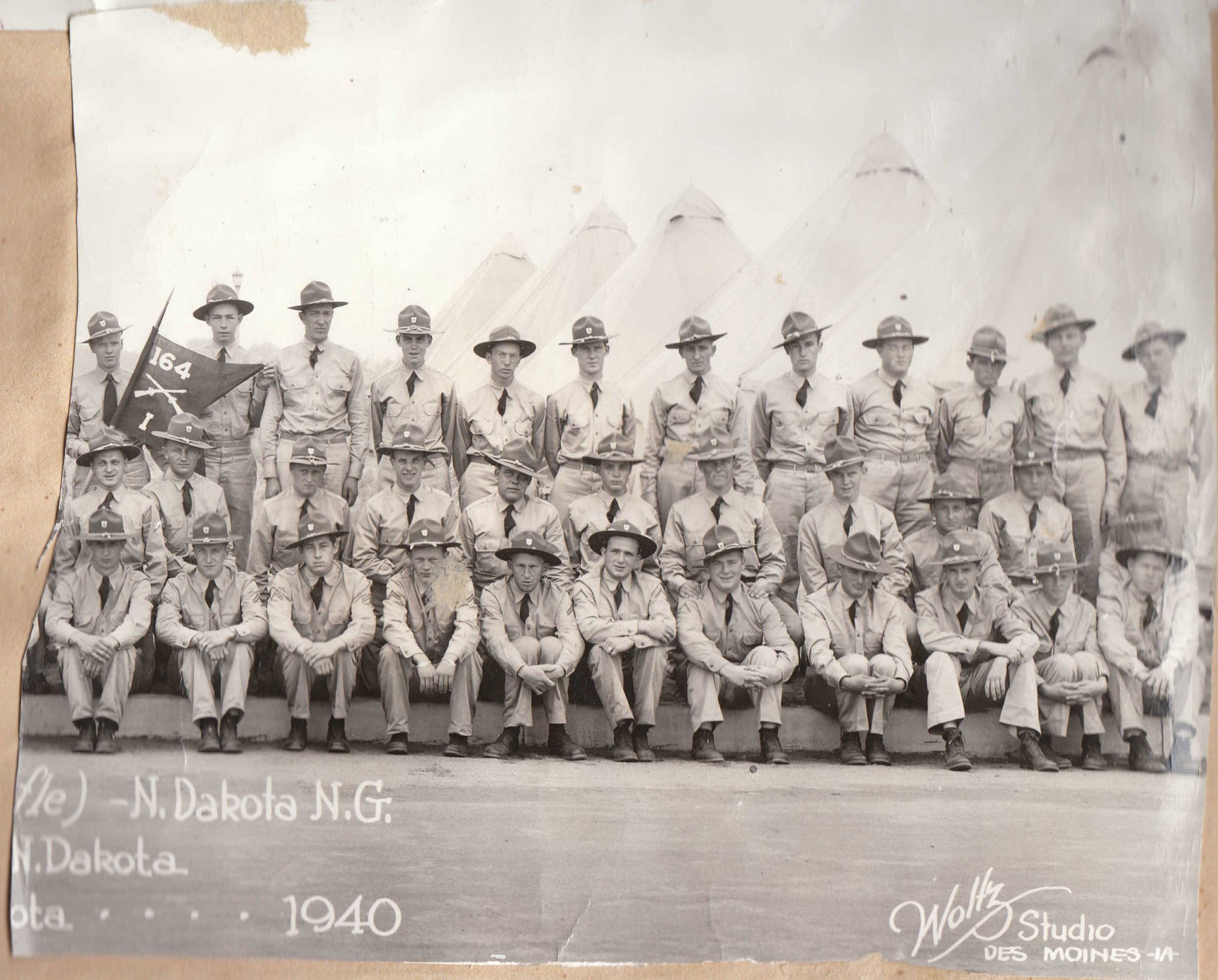 Black and white vintage team photo of young men in military uniforms, some with hats, posing outdoors with wing-shaped structures in the background. One man holds a flag with the number 164. Text at the bottom indicates the group is from North Dakota, N.G., in 1940, taken by Waltz Studio in Des Moines, Iowa.