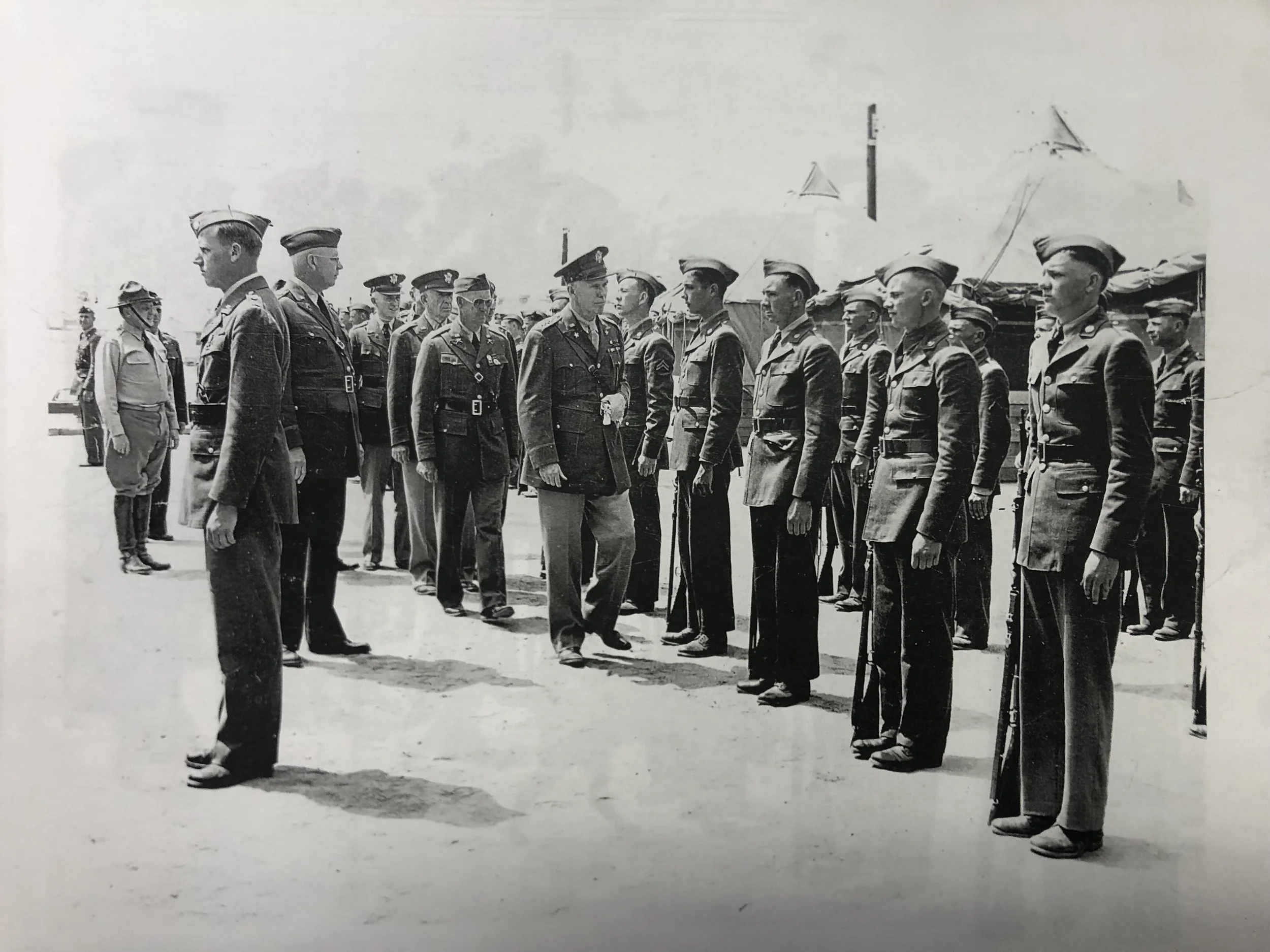 A group of military officers and personnel standing in formation in an outdoor setting, with a tent and flags in the background, during a formal ceremony or inspection.