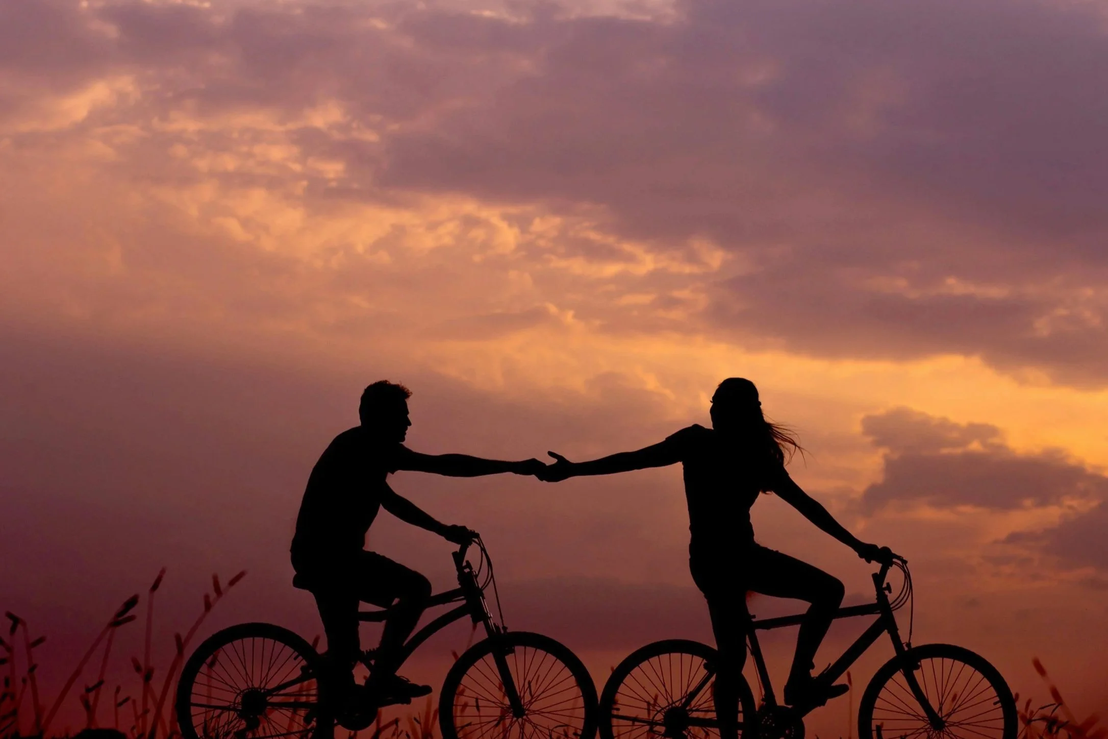 Silhouette of a young man and woman riding bicycles and reaching out to hold hands against a colorful sunset sky.