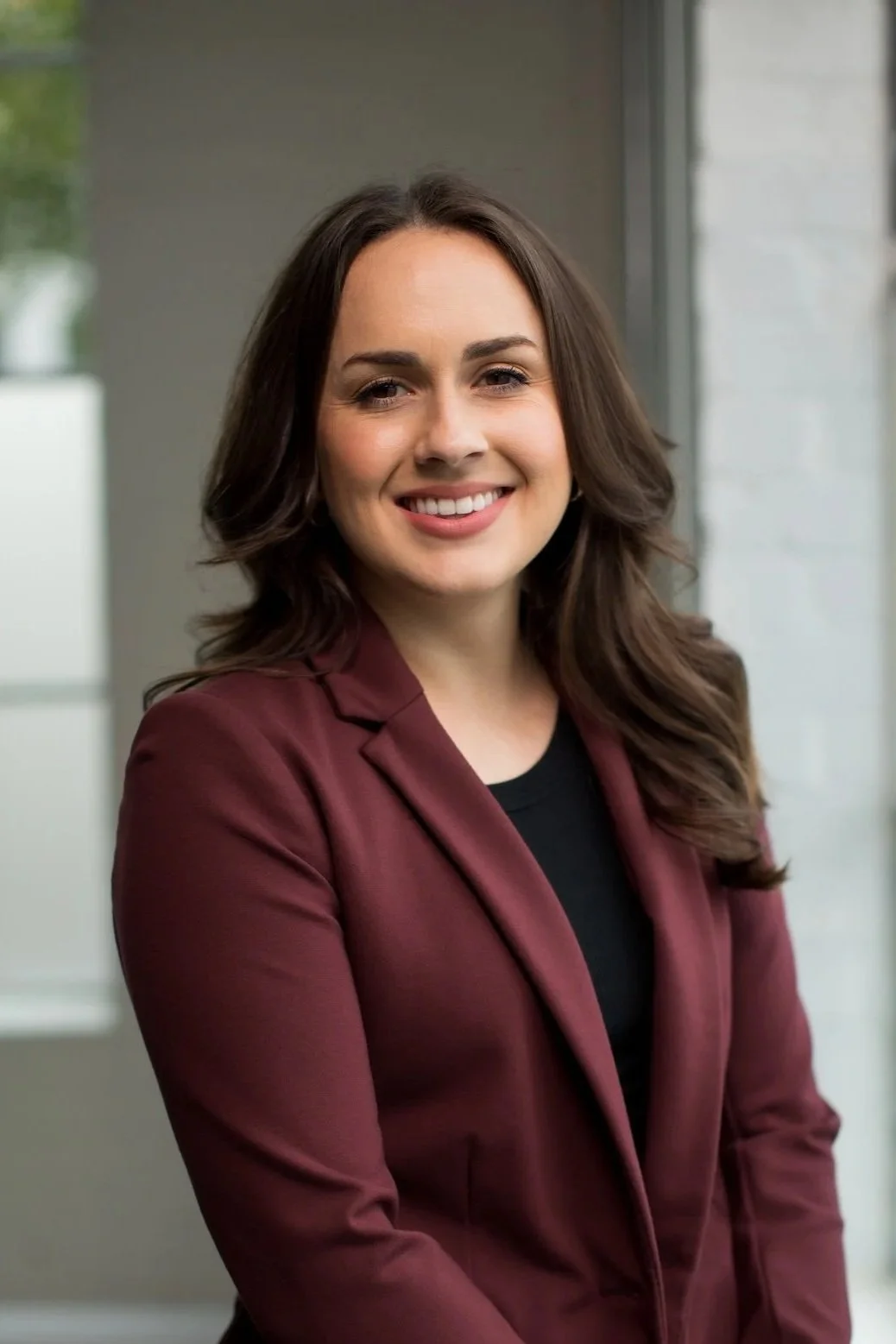 A woman with long brown hair wearing a maroon blazer and black top, smiling in front of a window.