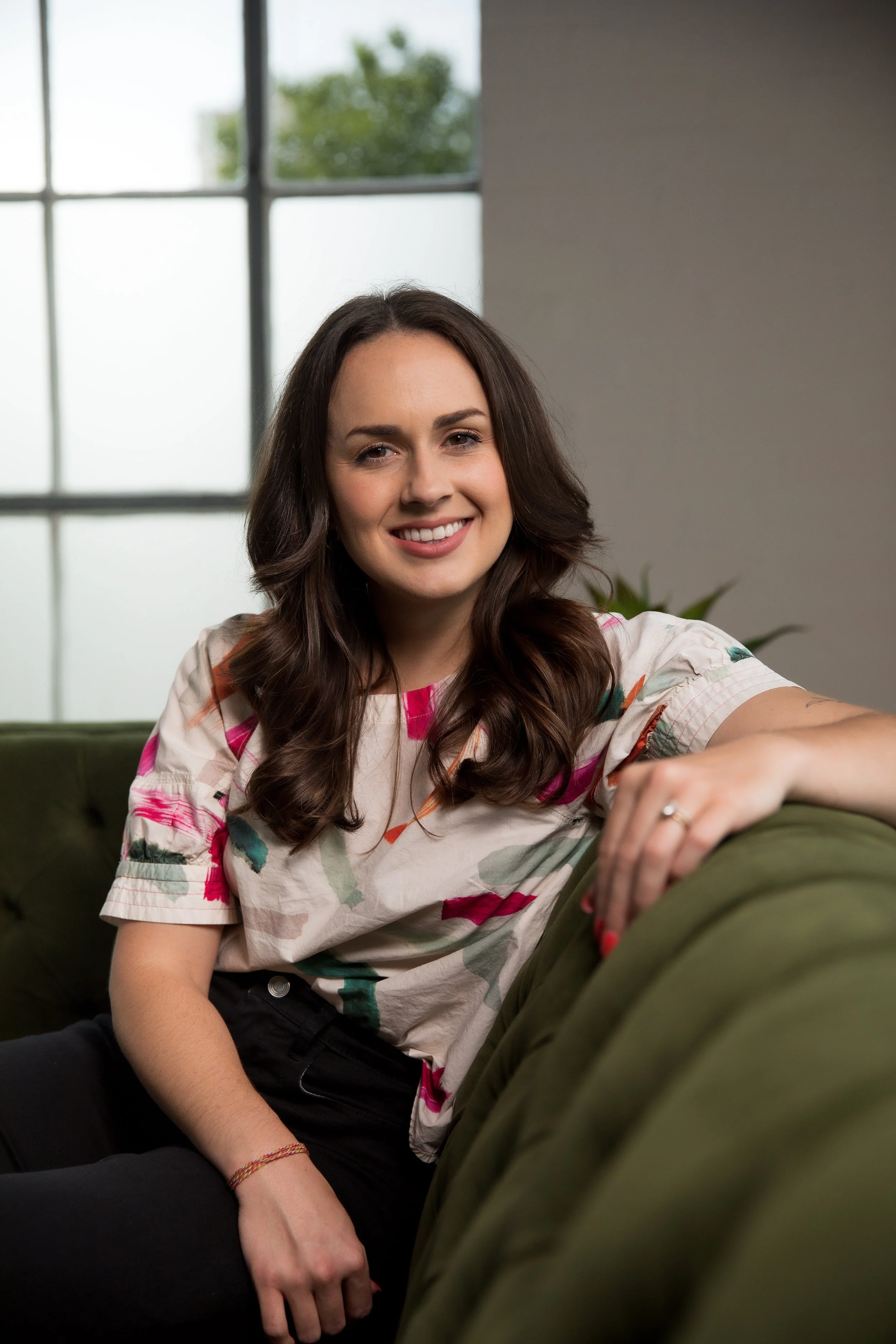 A young woman with long dark hair smiling while sitting on a green couch in a well-lit room with large windows and a gray wall in the background.
