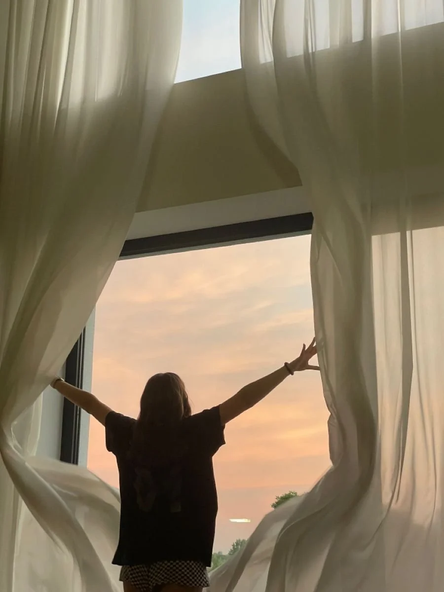 Person standing at open window with arms outstretched, looking at a colorful sunset sky, surrounded by sheer curtains.