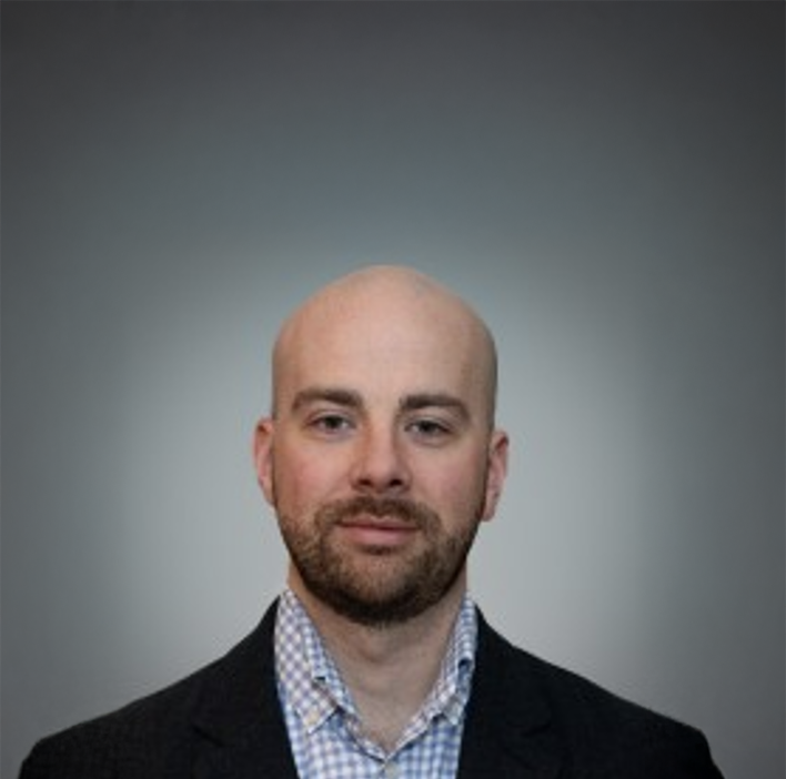 A portrait of a man with a shaved head, beard, wearing a checked shirt and blazer against a plain background.
