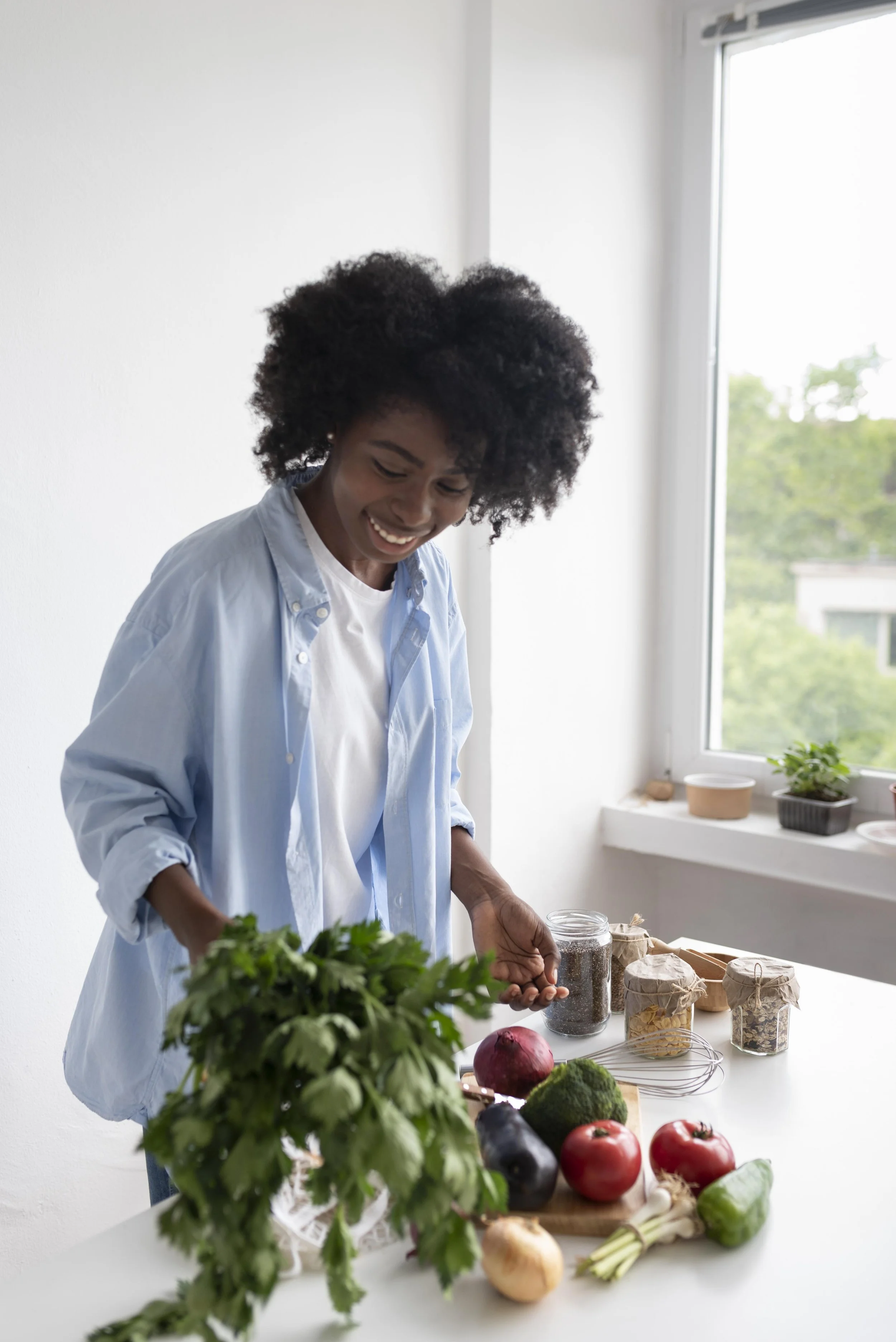 A woman with curly hair in a blue shirt and white t-shirt preparing vegetables and herbs on a kitchen counter, smiling, with a window showing greenery outside.