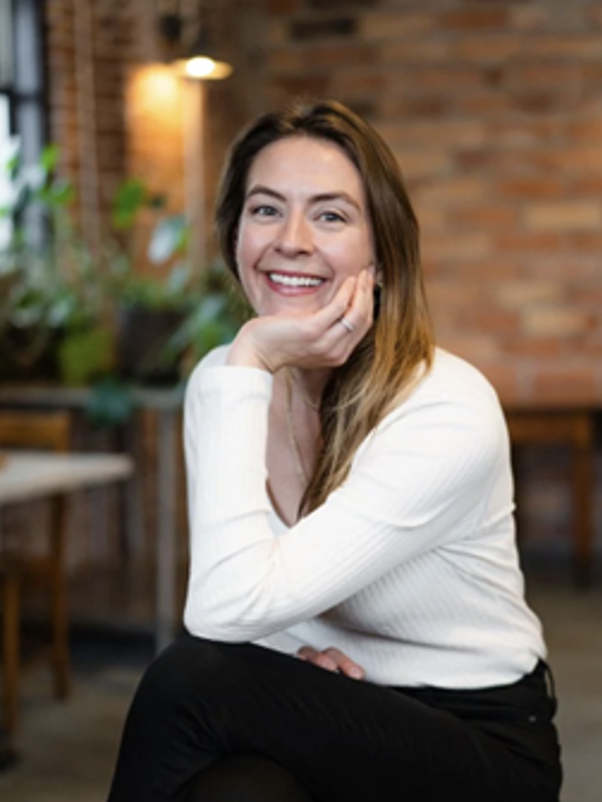 Smiling woman with long hair, sitting in a cozy restaurant or cafe with brick walls and potted plants in the background