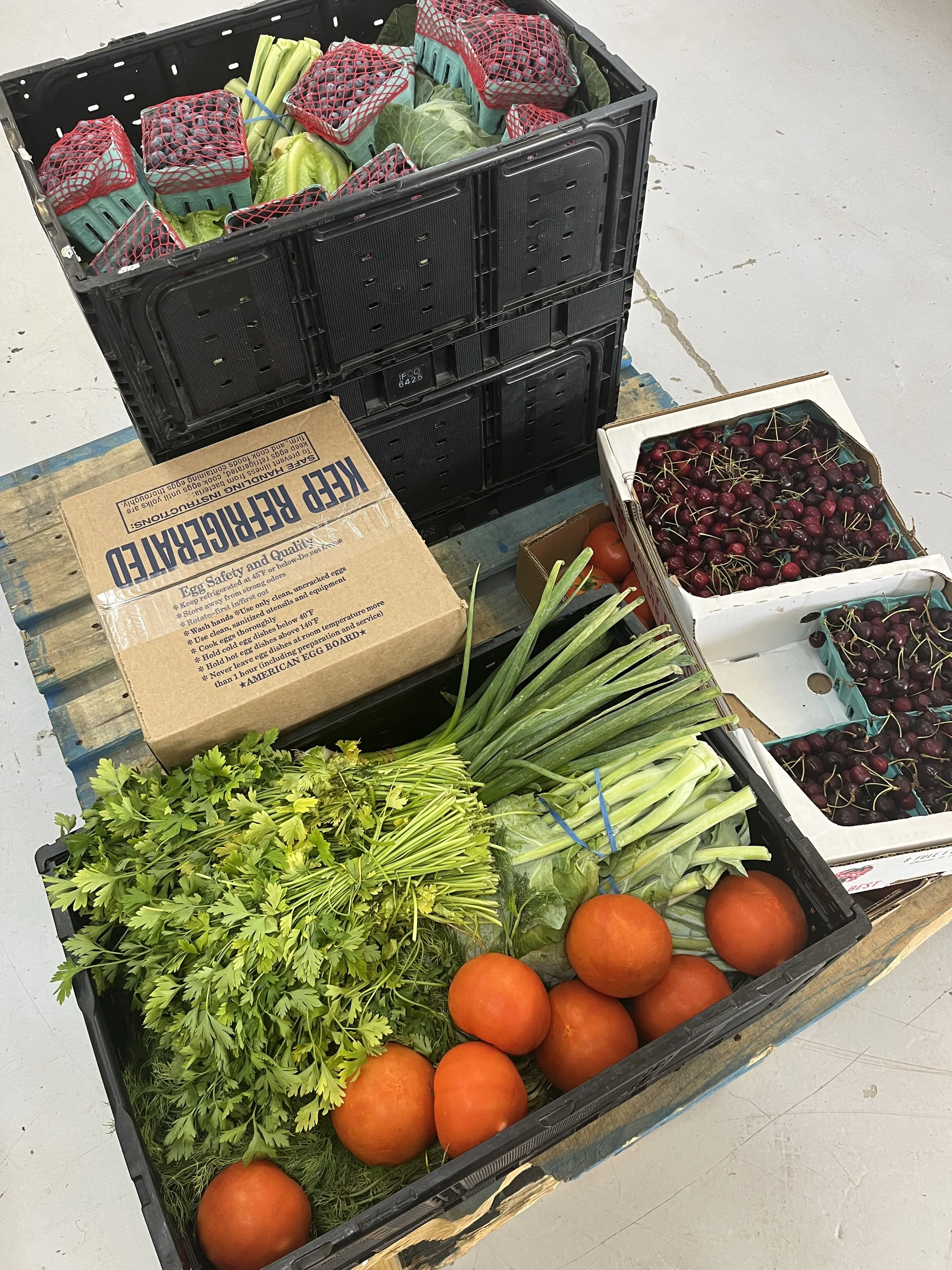 A black plastic crate filled with fresh vegetables, leafy greens, and tomatoes, placed on a wooden pallet alongside boxes of cherries and a cardboard box of egg cartons.