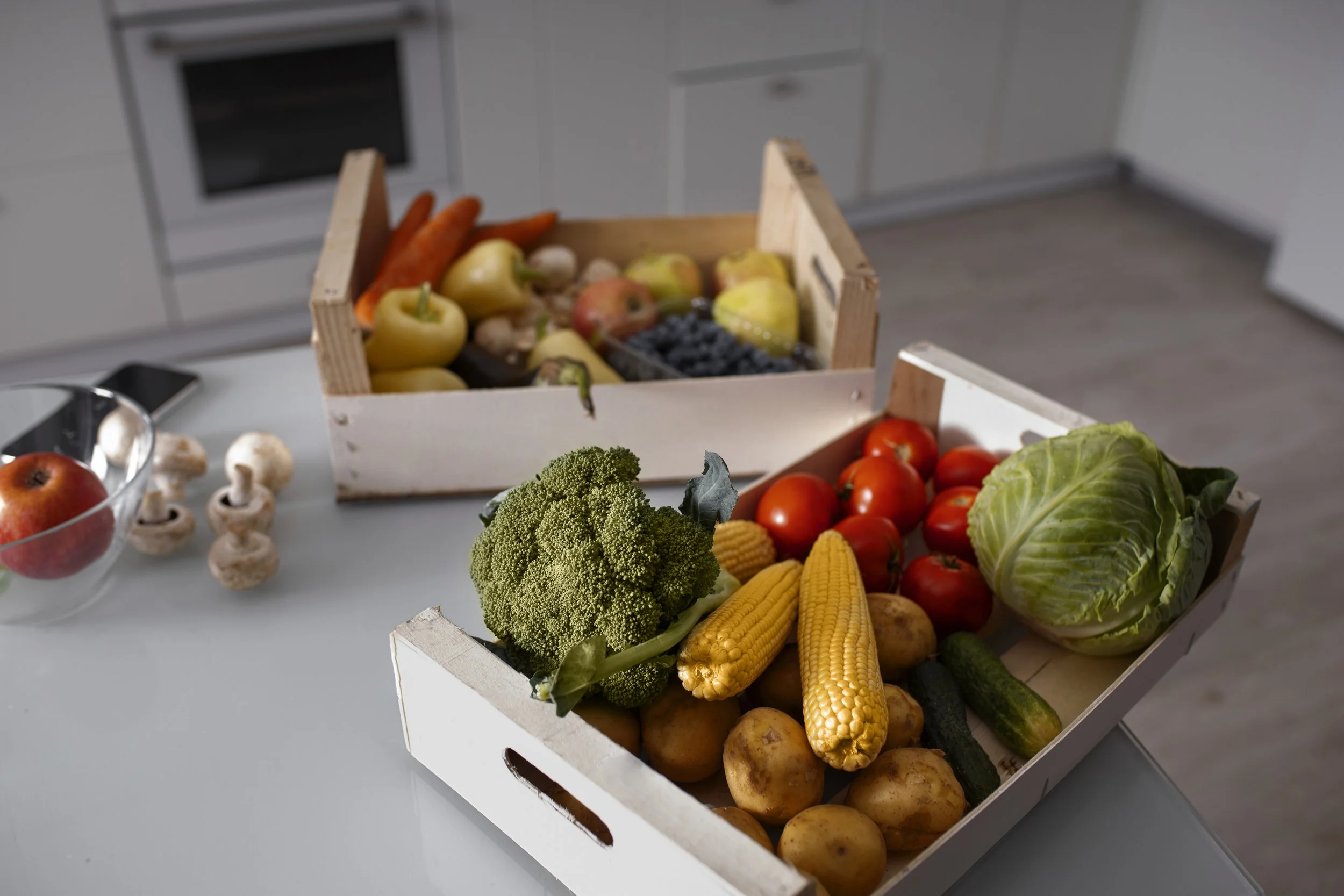 Fresh vegetables and fruits in white crates on a kitchen counter. Includes broccoli, corn, potatoes, cabbage, tomatoes, zucchini, apples, blueberries, mushrooms, and carrots.