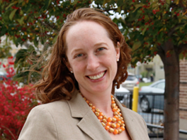 Portrait of a smiling woman with curly red hair, wearing a beige blazer and an orange beaded necklace, outdoors with trees and cars in the background.