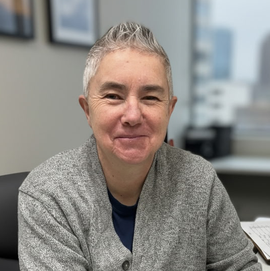A smiling woman with short gray hair sitting in an office with a blurred background.