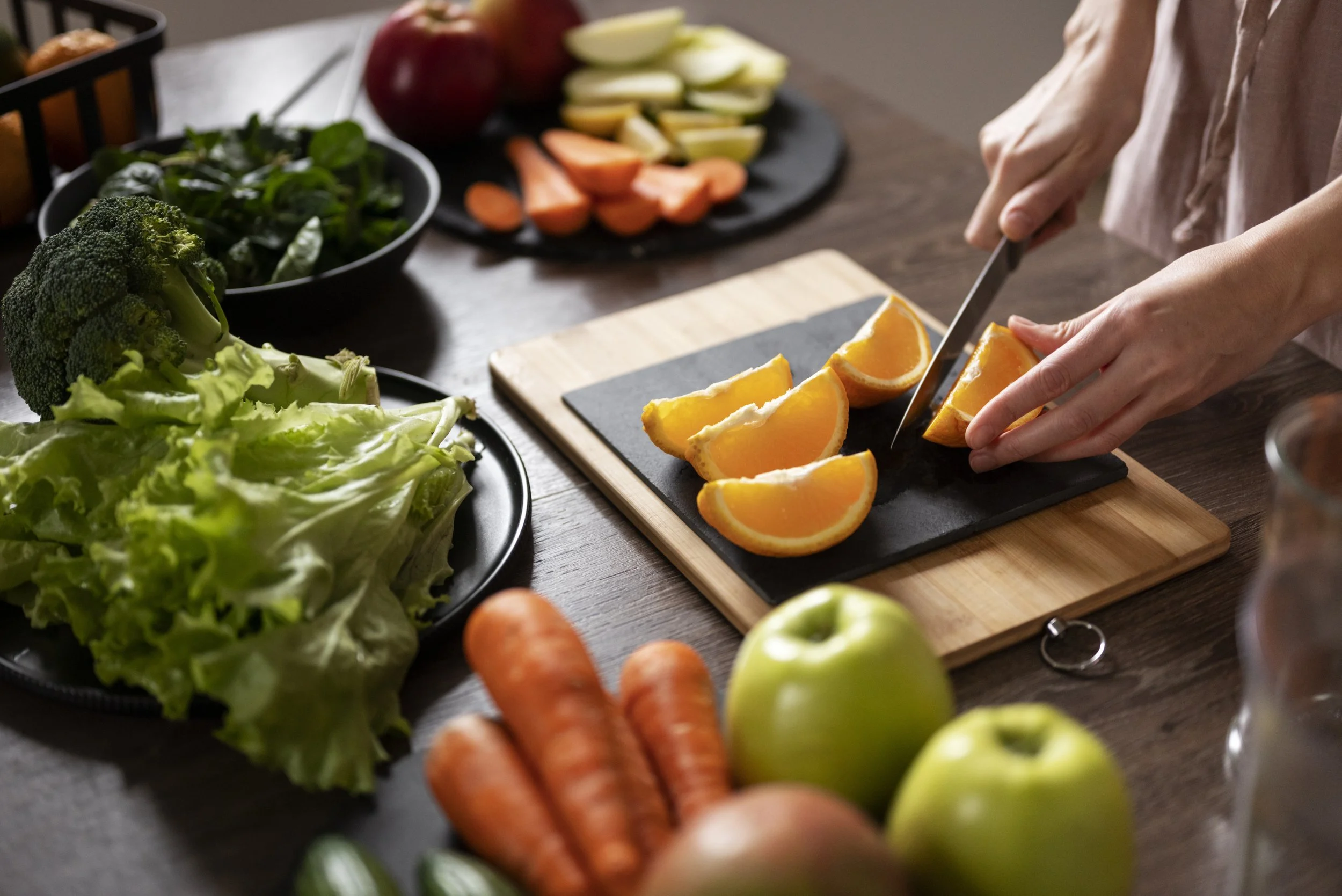 Person slicing orange wedges on a black cutting board in a kitchen with various fresh vegetables around, including carrots, apples, broccoli, lettuce, and tomatoes.