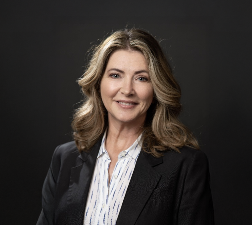 A professional woman with shoulder-length wavy blonde hair, wearing a black blazer and a white striped blouse, smiling against a dark gray background.
