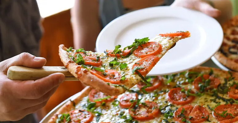 Someone serving a slice of pizza topped with tomatoes, green onions, and herbs from a large pizza.