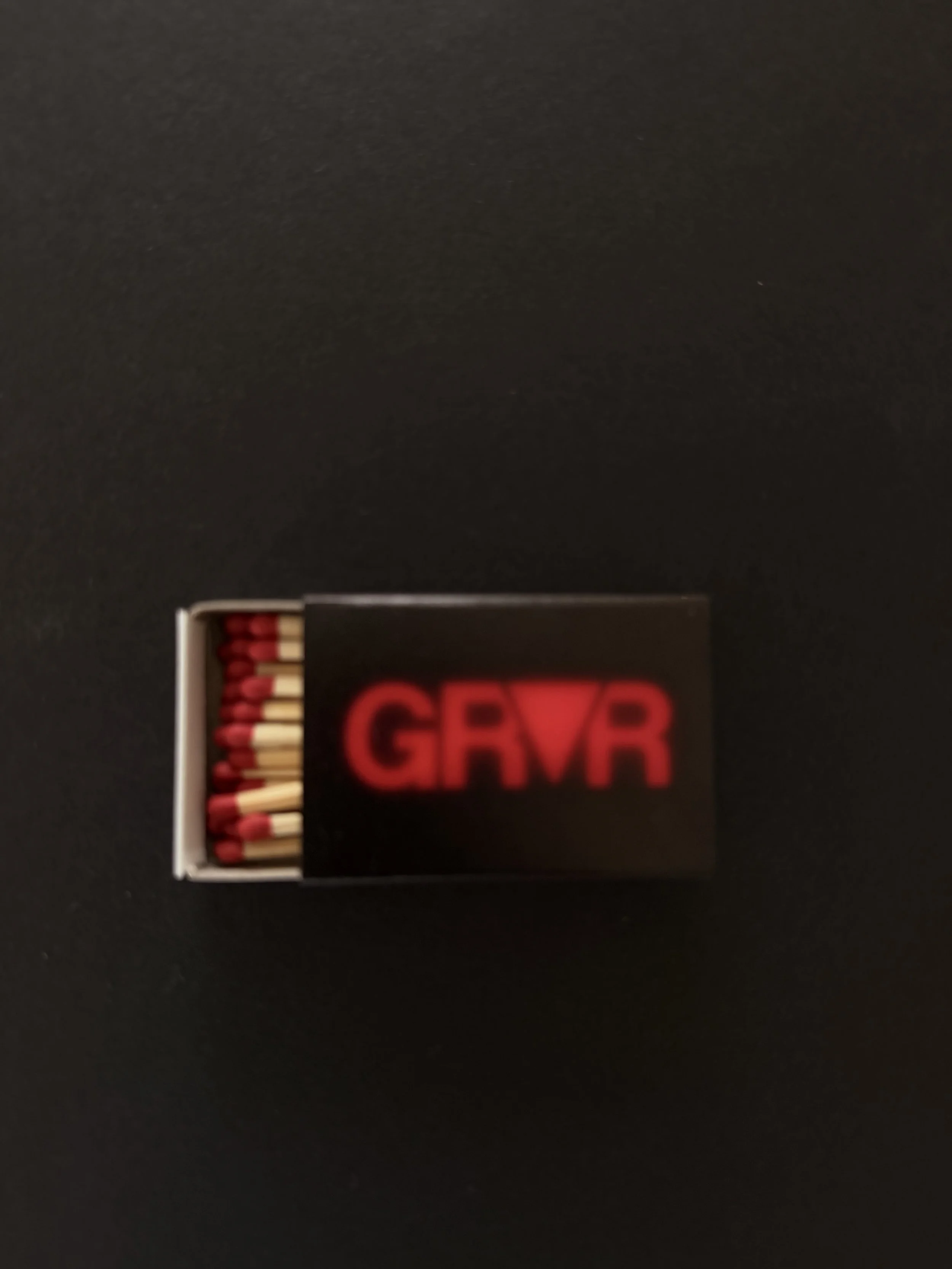 A box of matches with a black cover featuring red text and a triangle logo, on a dark surface.
