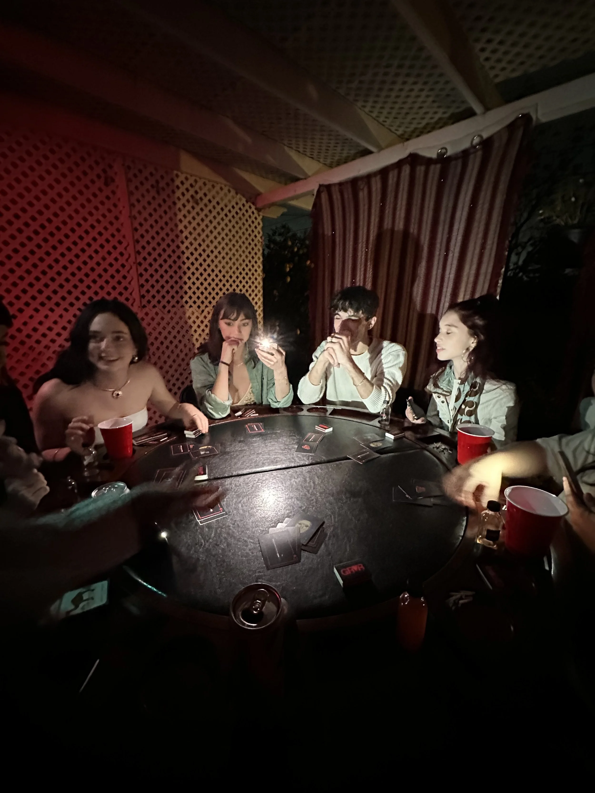 Group of people playing cards around a circular table at night, some holding drinks, with a dimly lit, decorated outdoor setting.