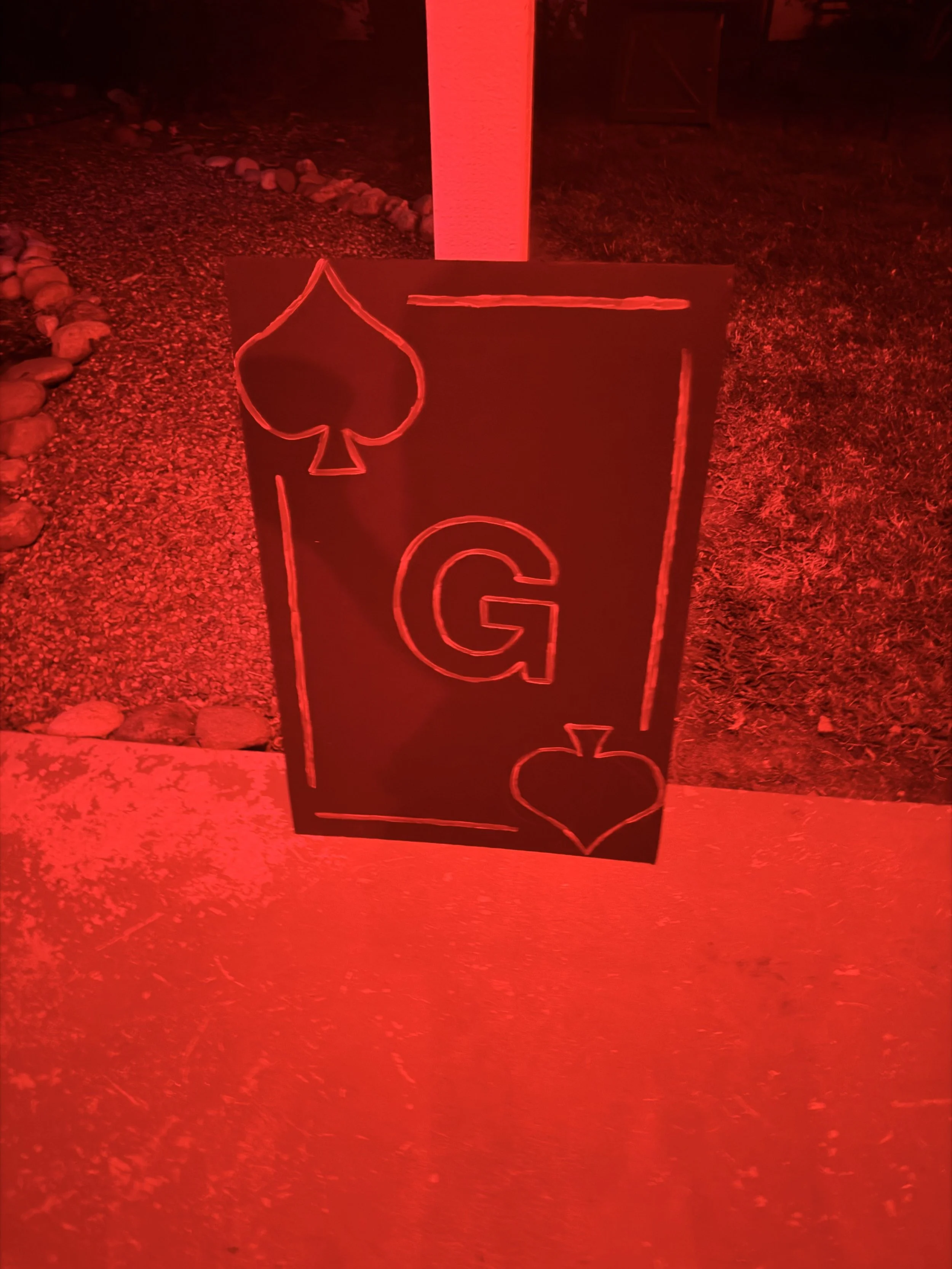 A black sign with a large red letter G and red outlines of playing card suits, a spade and a heart, illuminated by red lighting outside on a sidewalk near a grassy area and decorative rocks.