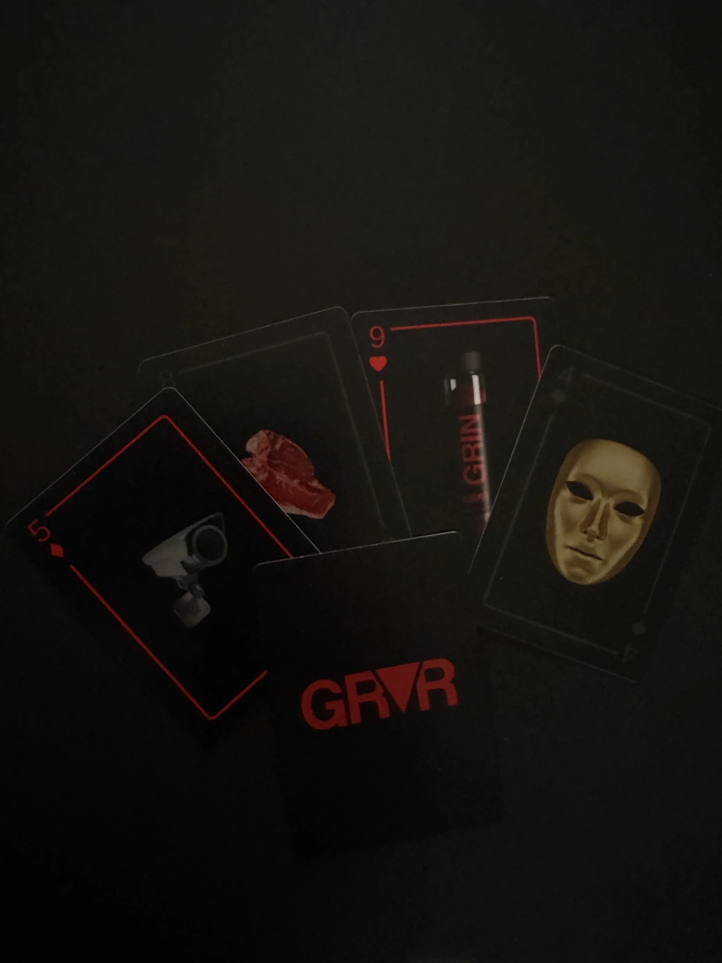 Dark background with four playing cards, including a 5 of diamonds featuring a gun, a 9 of hearts with a spray bottle labeled 'GRAIN', an unknown card, and a card with a gold mask. The deck box labeled 'GRVR' is also visible.