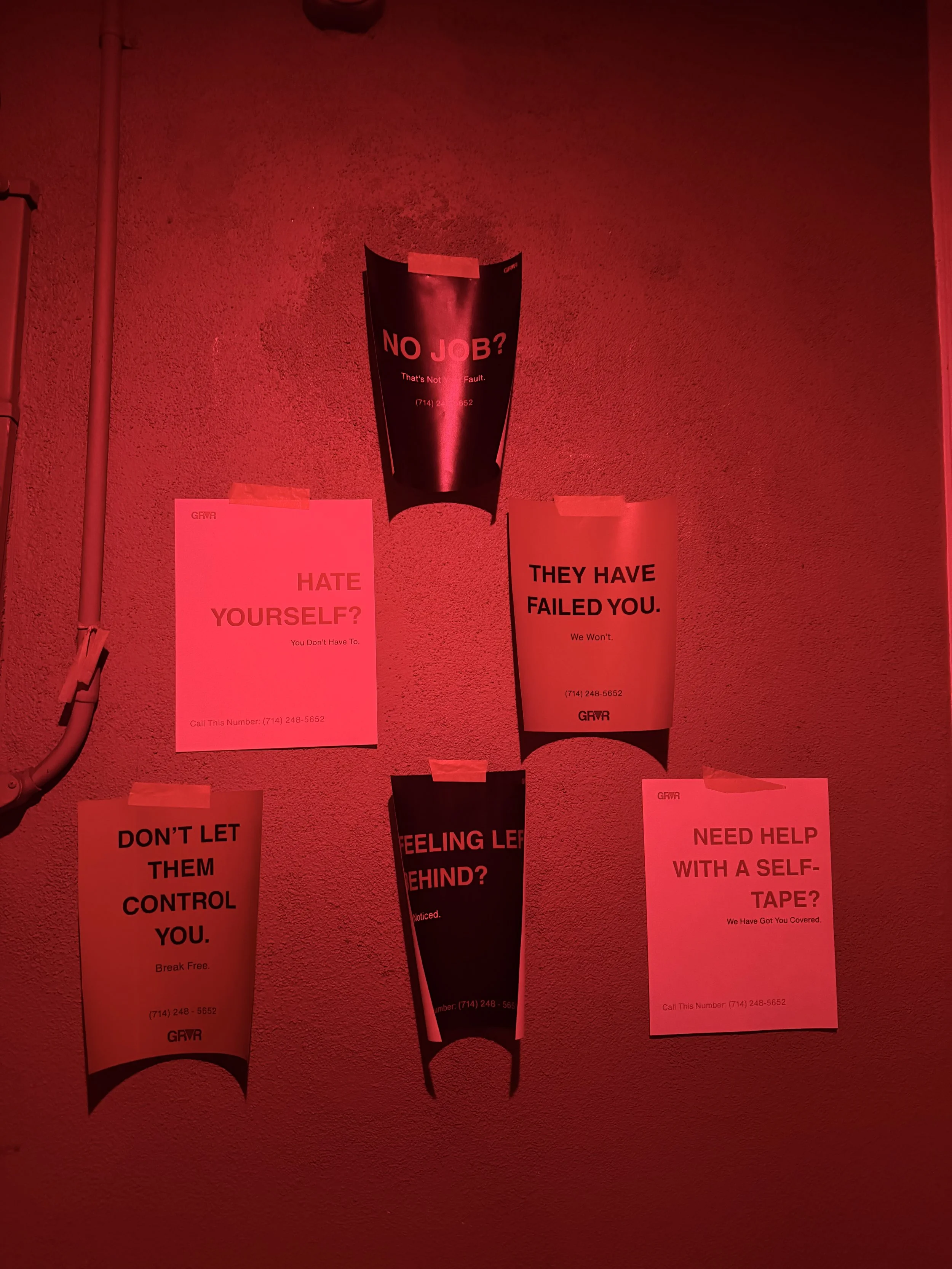 Several flyers on a red-lit wall with motivational messages including 'No Job? That's Not Your Fault.' 'Hate Yourself? You Don't Have To.' 'They Have Failed You.' 'Feeling Left Behind?' 'Don't Let Them Control You.' and 'Need Help With a Self-Tape? We Have Got You Covered.'