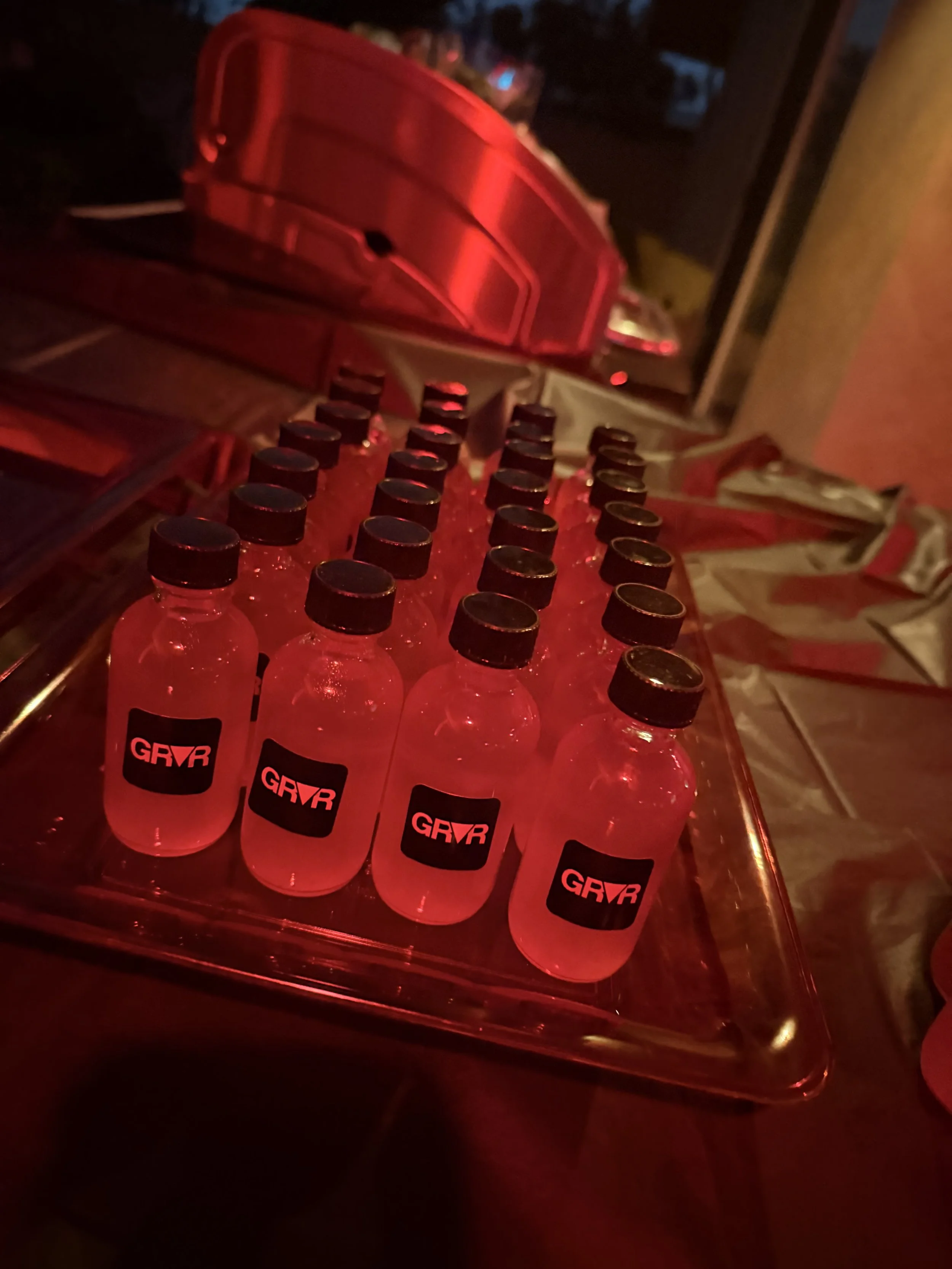 Miniature bottles of pink-colored shot liquor with black caps and black labels that have red text saying 'GRVR' are arranged on a silver serving tray at a social event or party.