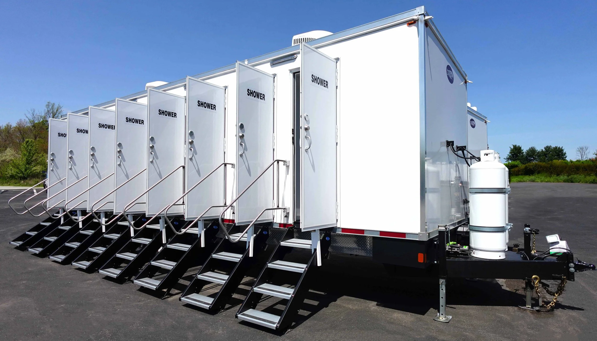 A mobile shower trailer with multiple individual shower stalls, each equipped with a door, steps, and handrails, situated outdoors on a paved surface under a clear blue sky.