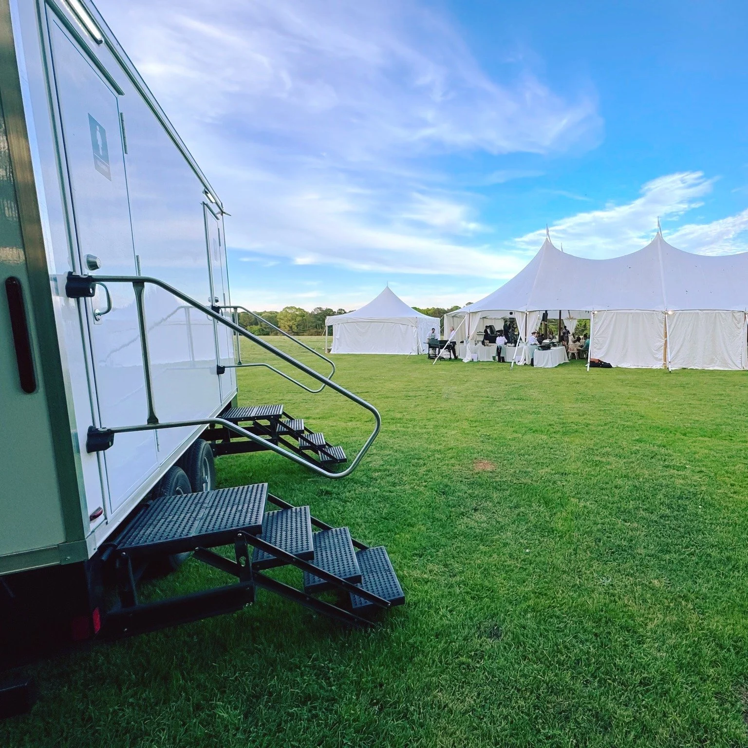 A mobile trailer with metal stairs leading up to its door, set on a grassy field near white event tents with pointed peaks. The sky is partly cloudy with blue and white clouds, and a group of people is gathered under the tents.