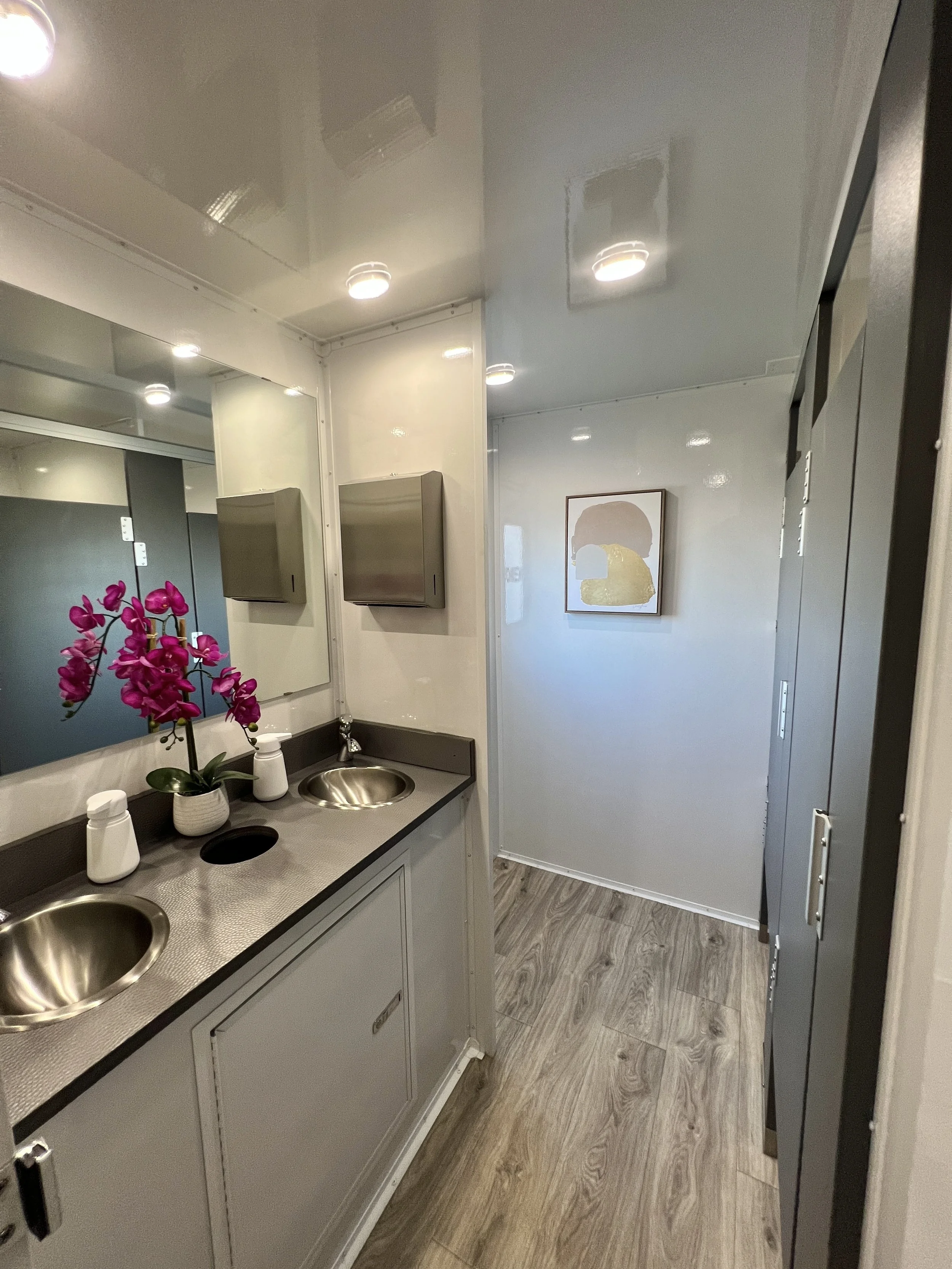 A small bathroom or break room with dual stainless steel sinks, a potted purple orchid on the counter, mirror, and wall-mounted paper towel dispensers in a mobile restroom trailer.
