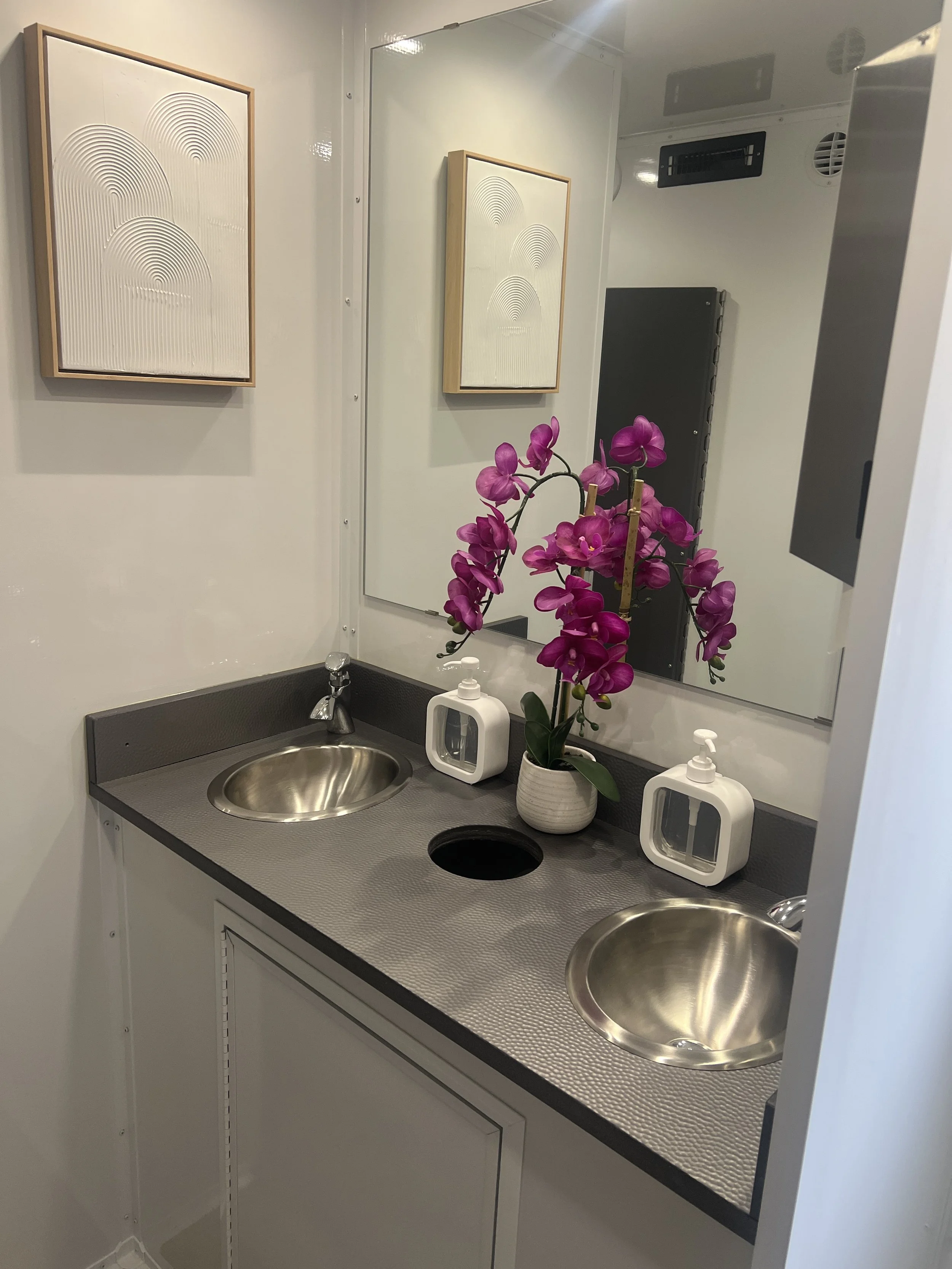 A bathroom with a dual sink vanity, a large mirror, purple orchid in a white pot, and two white soap dispensers. Two framed abstract art pieces hang on the wall.