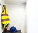 A safety vest hanging on a hook and a blue helmet on a small table in a plain, white-walled room in a bunkhouse rental trailer.
