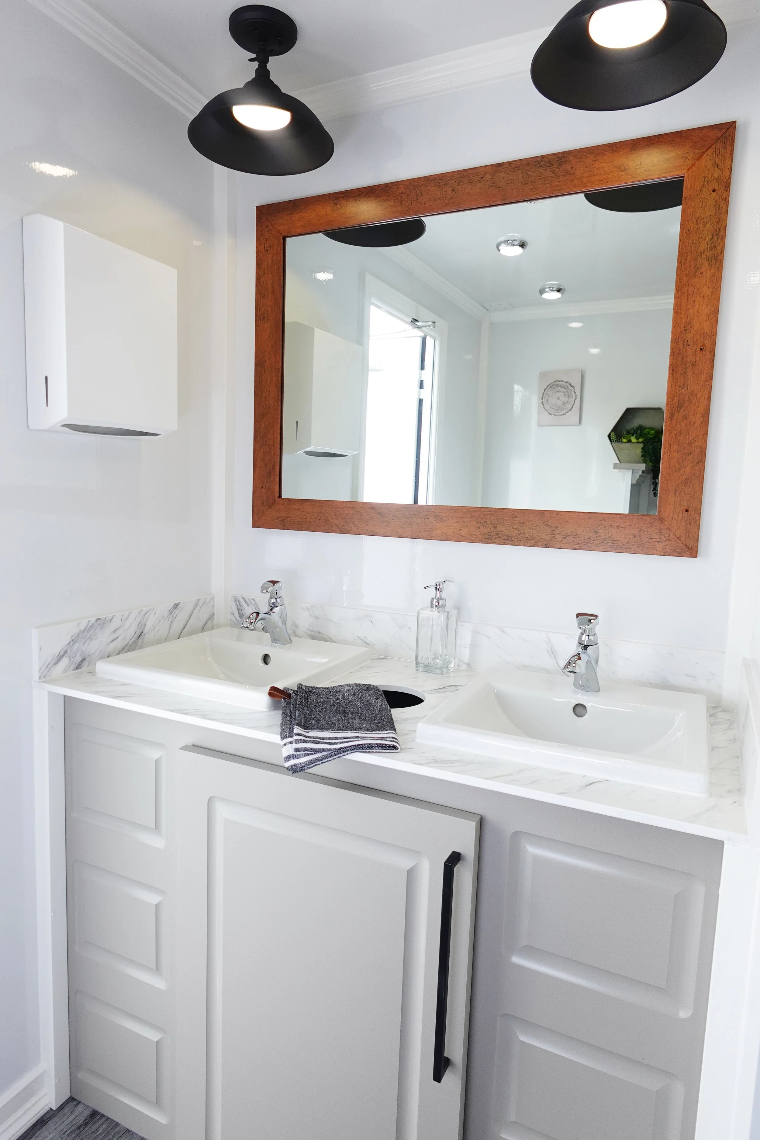 Bathroom double vanity with white cabinets, marble countertop, two sinks, and a large wooden-framed mirror. There is a black soap dispenser and a gray towel on the countertop. Interior of a mobile restroom trailer.