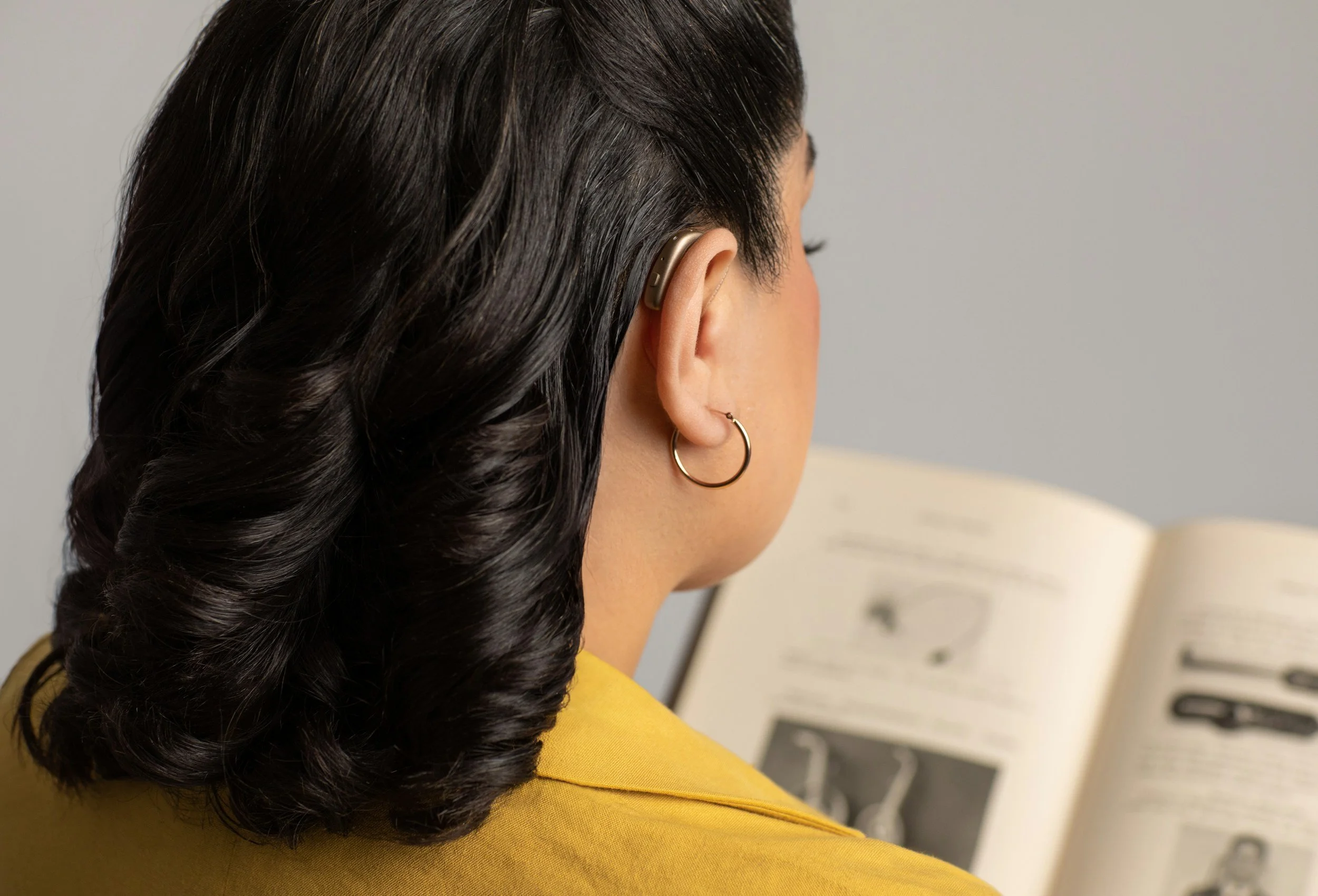 A woman with shoulder-length black hair wearing a yellow top, gold hoop earrings, and an earpiece, reading a book with text and illustrations.