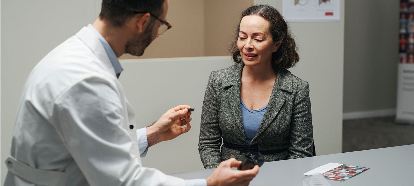 A doctor showing a patient a small object, possibly a medical device or sample, during a consultation in a clinic or hospital reception area.