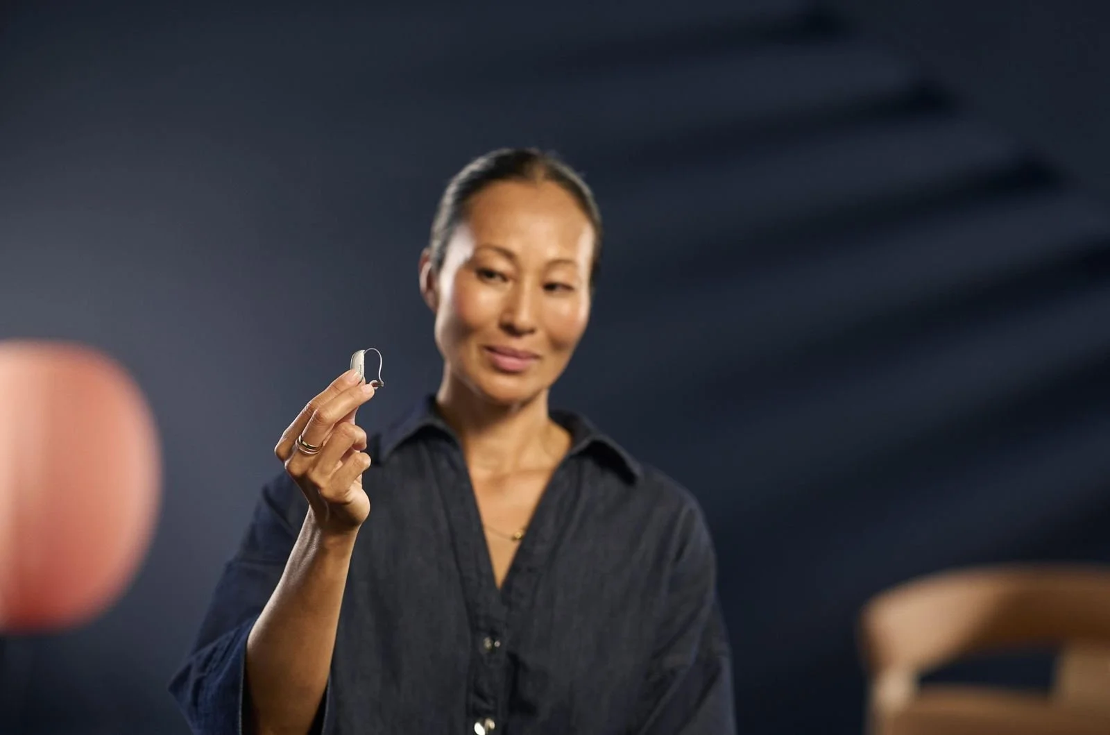 A woman holding a hearing aid with a thoughtful expression, wearing a dark shirt, against a dark background.