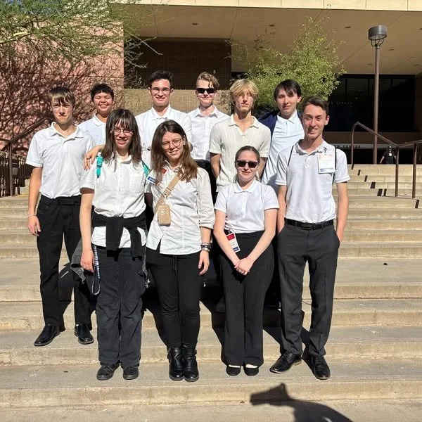 Group of students standing on stairs outside a building, wearing white shirts, with some holding bags or notebooks.