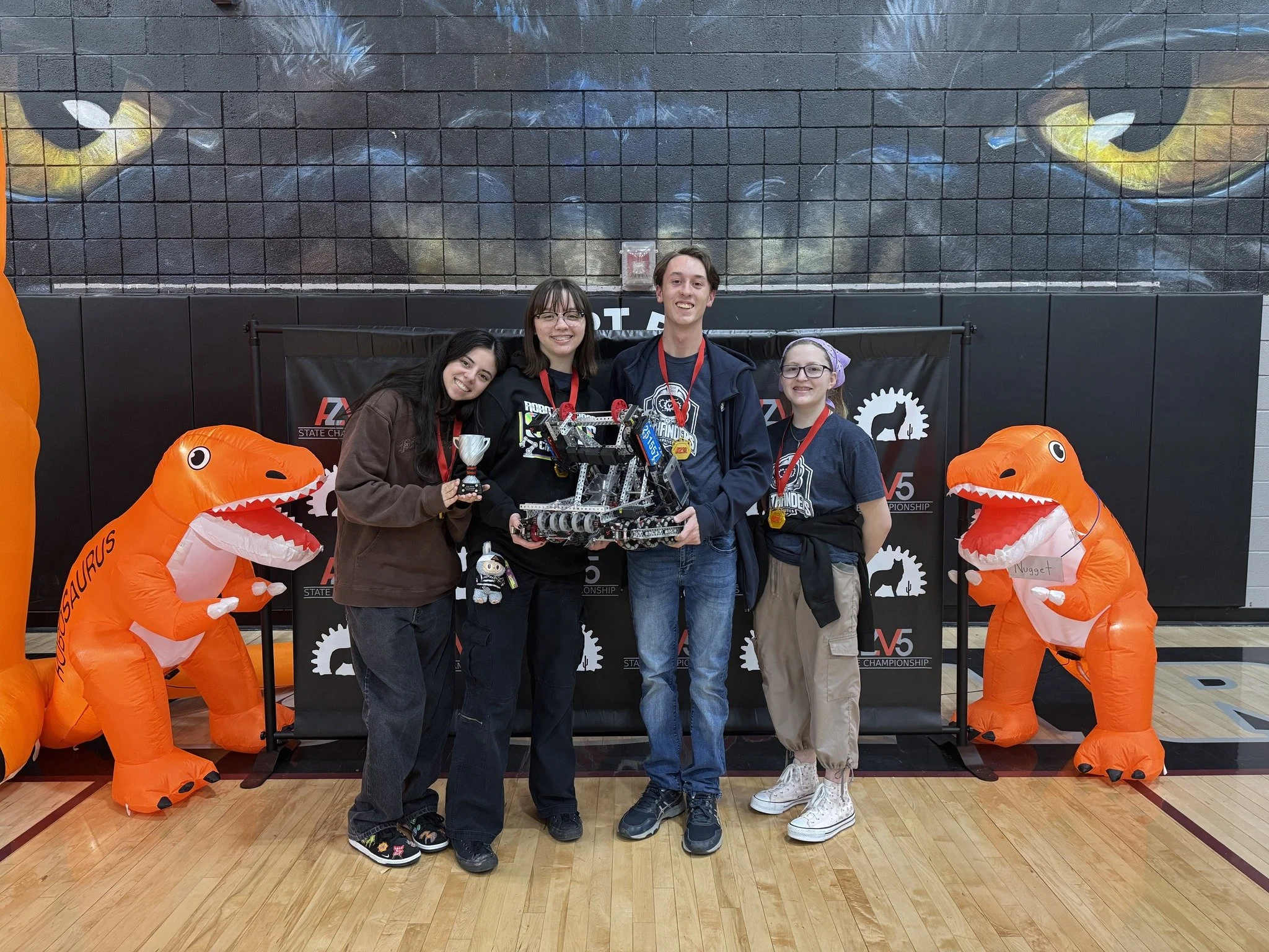 Four people standing together on a basketball court, holding a robot, with two large inflatable orange dinosaur figures on either side. They are celebrating their achievement at a robotics competition.