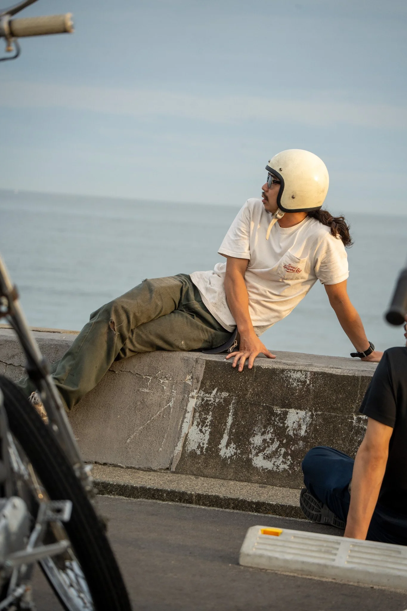 A person wearing a white helmet, beige t-shirt, and green pants is sitting on a concrete barrier near the water, looking to their left. Another person, partly visible, is sitting on the ground nearby, with a motorcycle in the foreground.