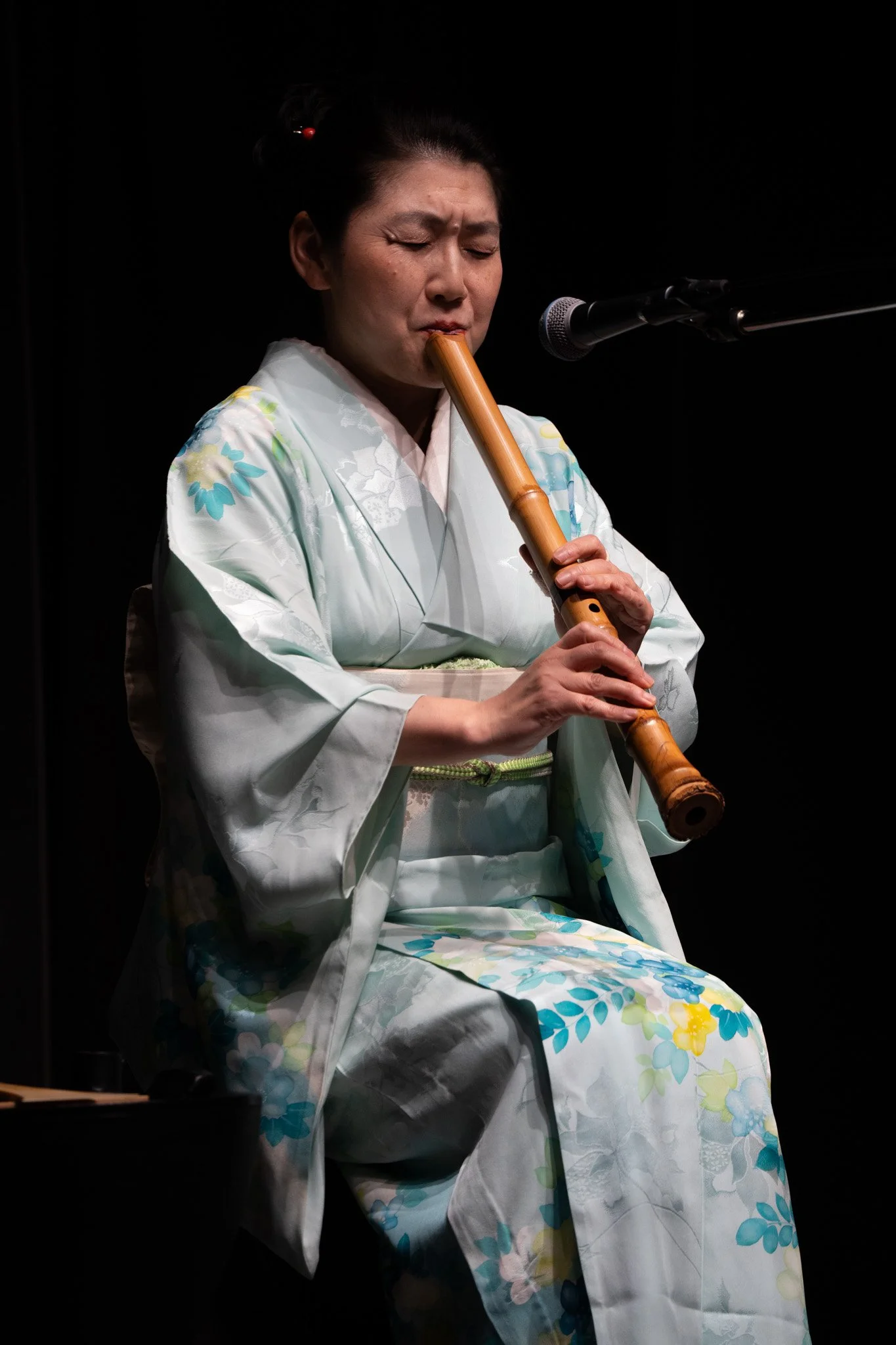 A woman in a light-colored kimono playing a bamboo flute on stage.