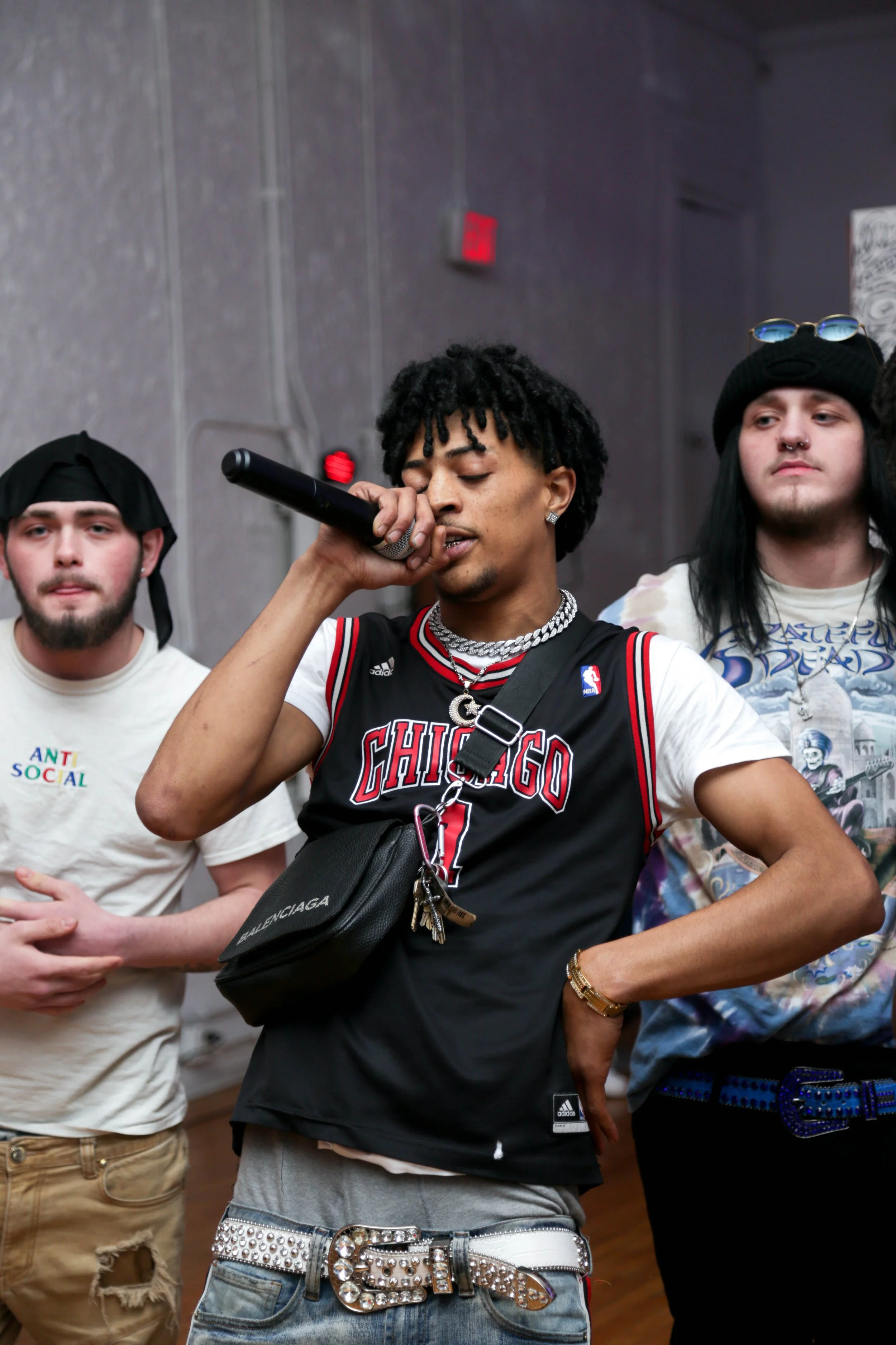 Young man with dreadlocks wearing a Chicago Bulls jersey, jewelry, and a cross necklace, holding a microphone and performing on stage with two other men in casual clothing.