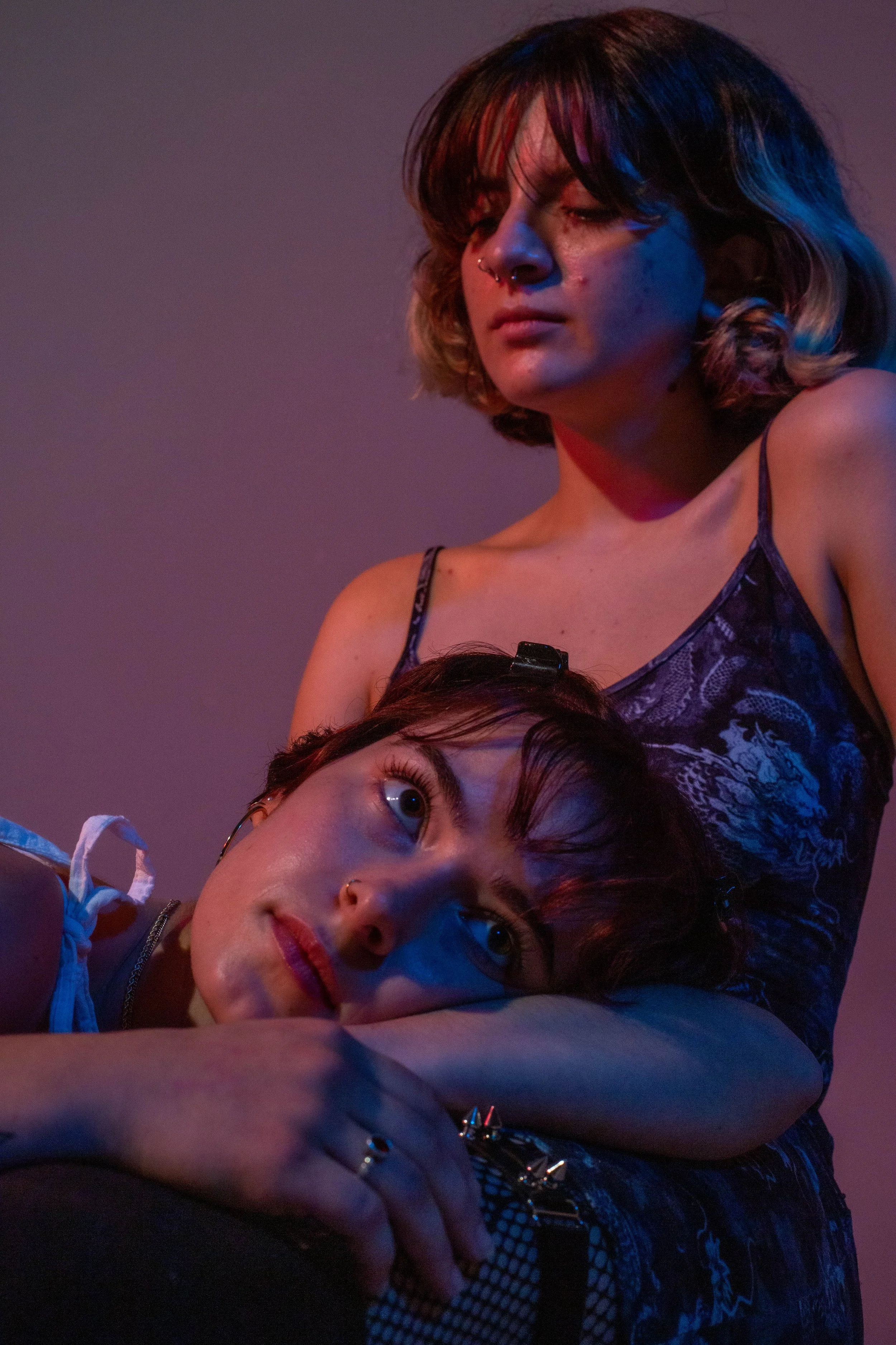 Two young women, one standing and one lying down with her head on the other woman’s arm, in a casual setting, illuminated by colorful, moody lighting.