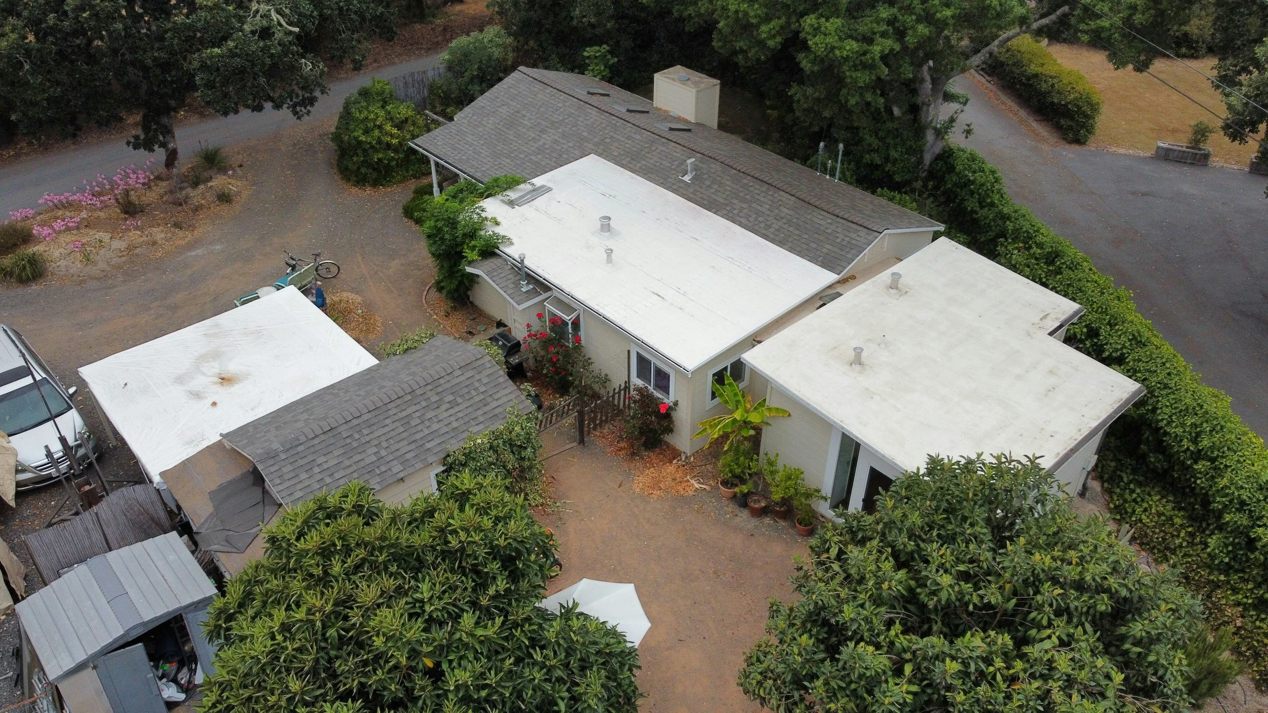 Aerial view of a residential area showing several houses with flat roofs, surrounded by trees and bushes, with a dirt driveway and parked vehicles.