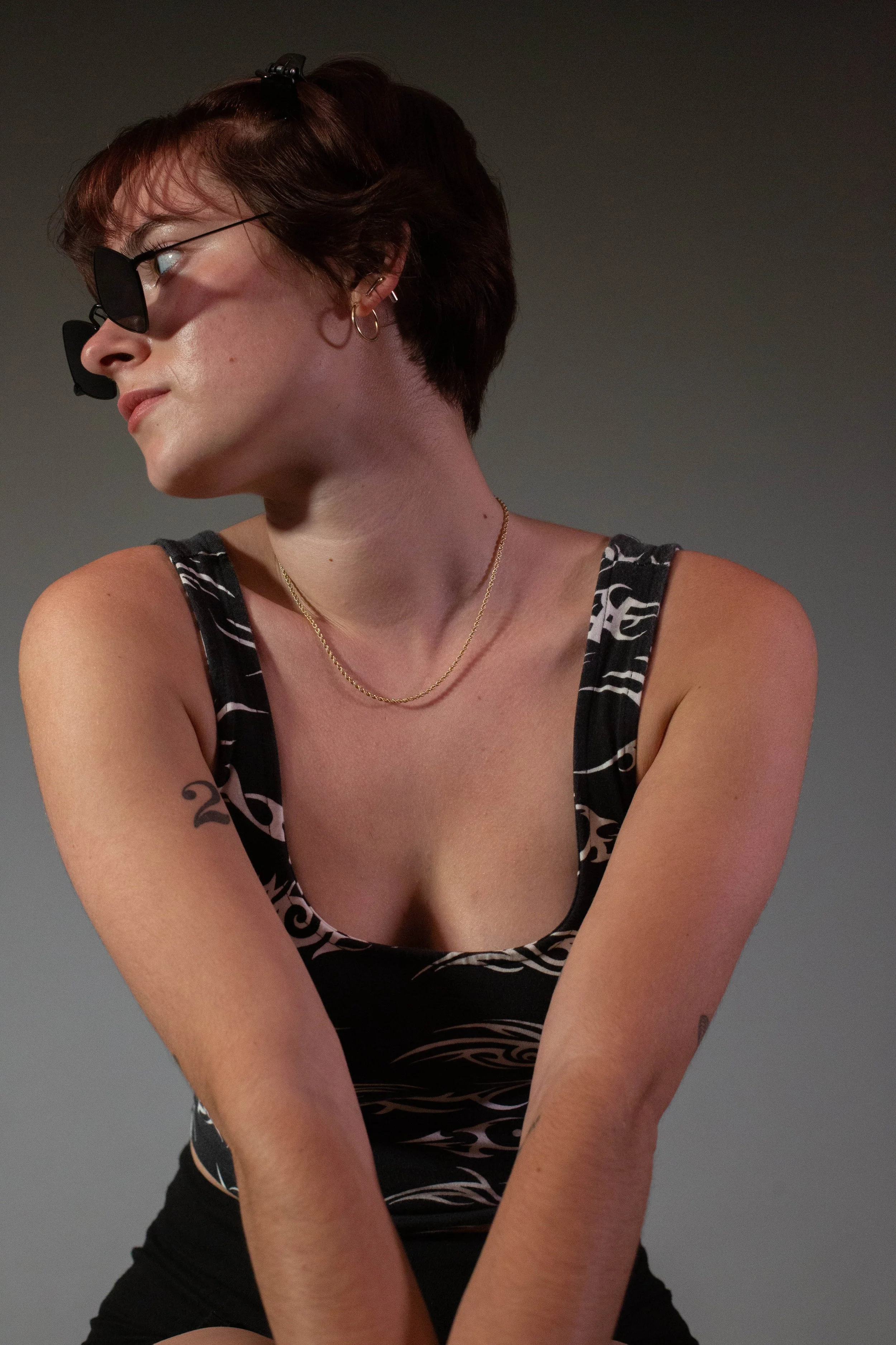 A woman with short brown hair, wearing sunglasses, hoop earrings, a gold chain necklace, and a black sleeveless top with white pattern, looking to her left against a plain background.
