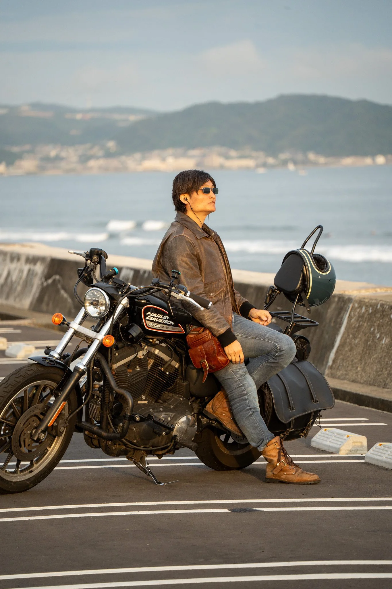 A person sitting on a Harley-Davidson motorcycle parked in a seaside parking lot, wearing sunglasses and a brown leather jacket, with mountains and ocean in the background.