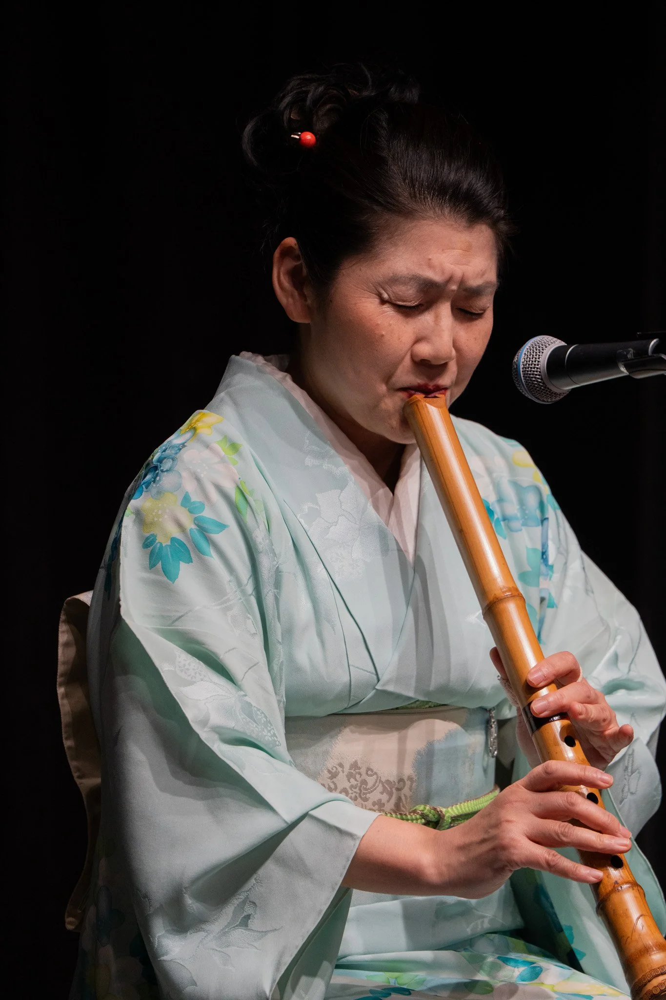 A woman dressed in traditional Japanese attire playing a bamboo flute in front of a microphone.