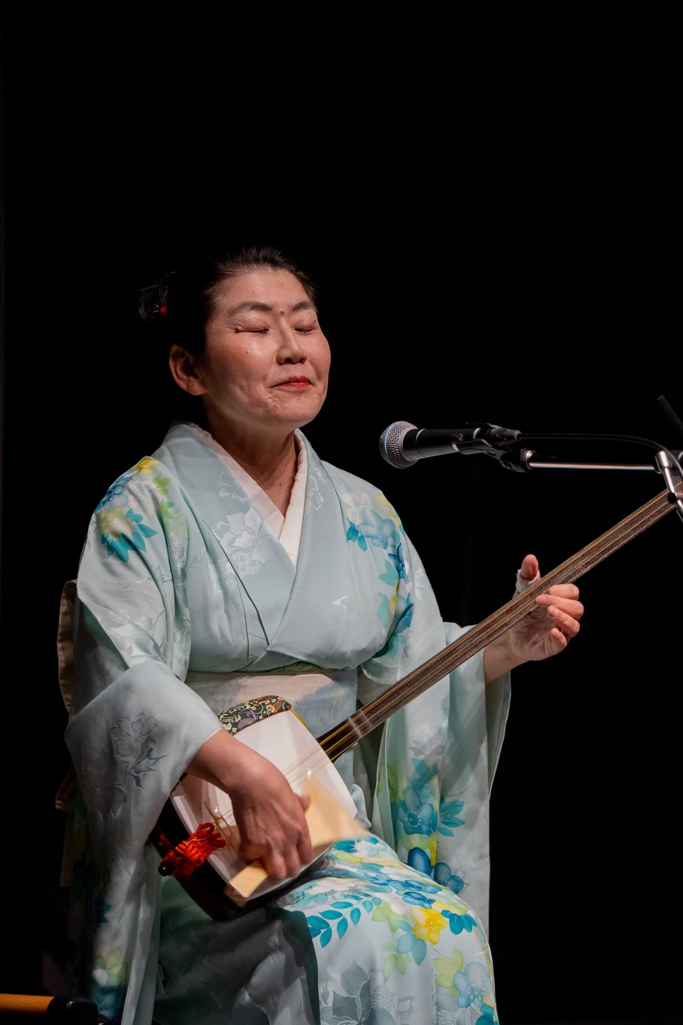 A Japanese woman in traditional kimono playing a shamisen on stage, with closed eyes and a gentle expression.