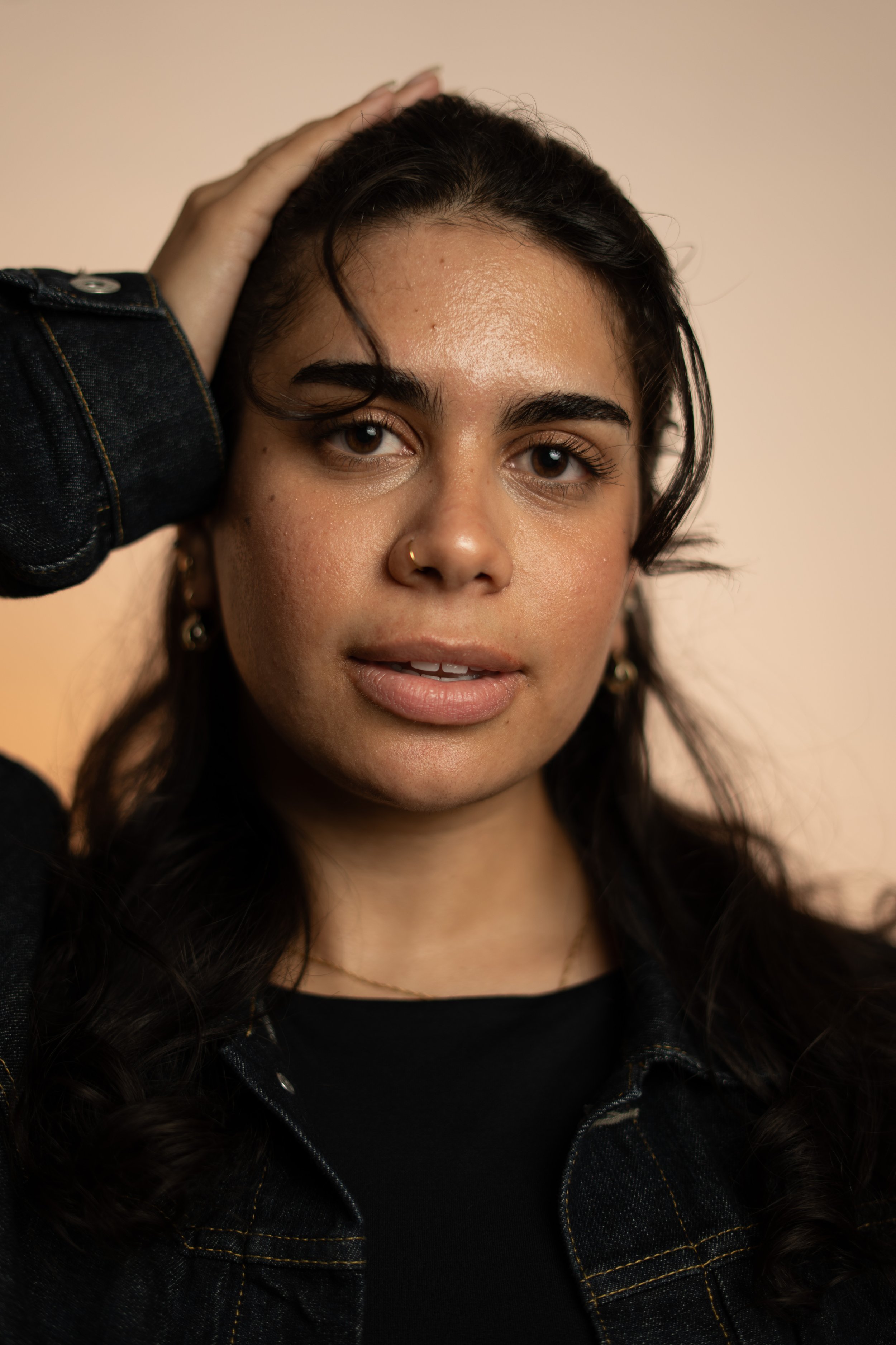 A young woman with dark hair, wearing a black top and jacket, touching her head with her hand, and looking directly at the camera.