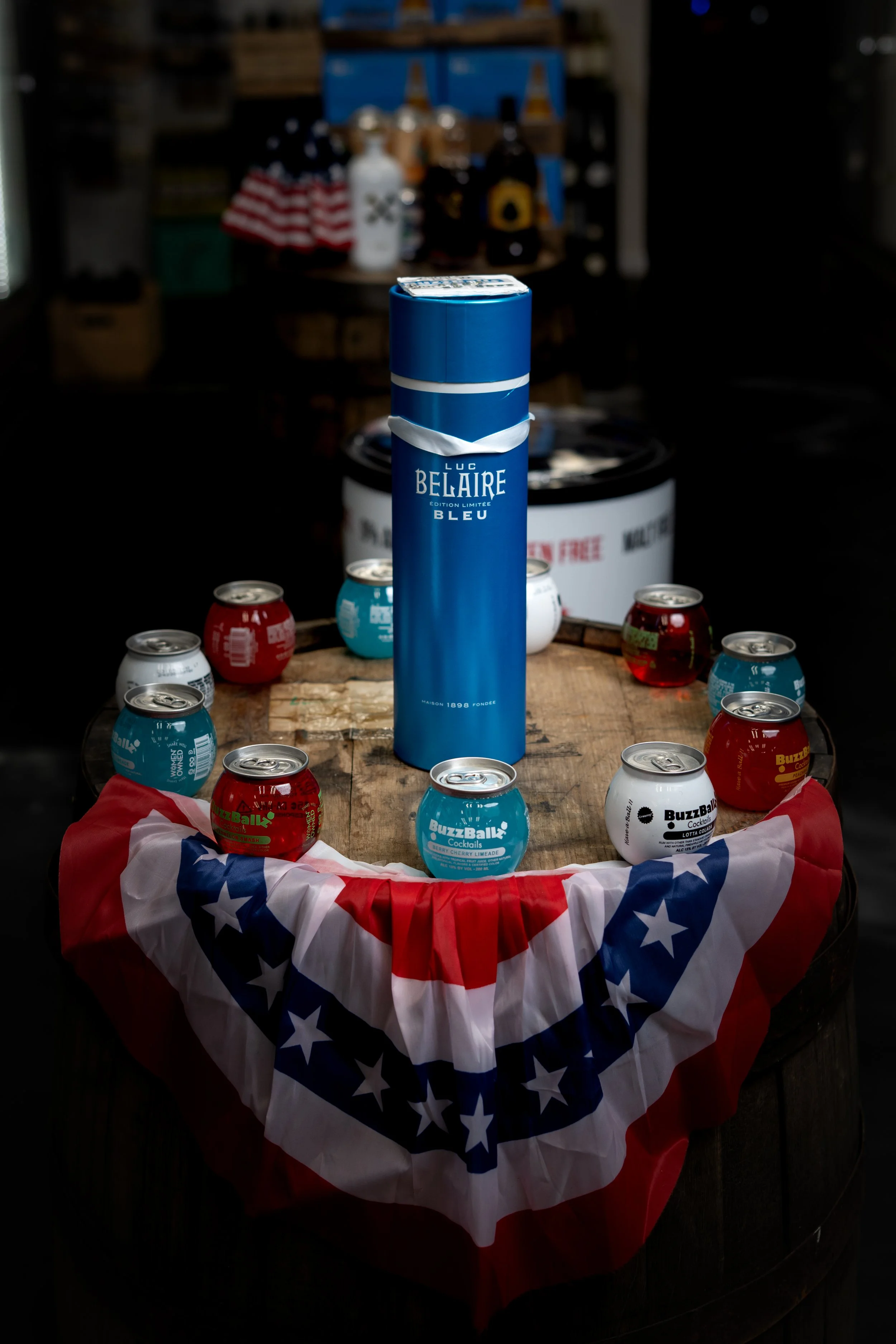 A wooden table decorated with a patriotic red, white, and blue bunting and surrounded by cans of BuzzBallz cocktails, with a tall blue box of Luc Belaire Bleu champagne in the center, set in a dark indoor room.