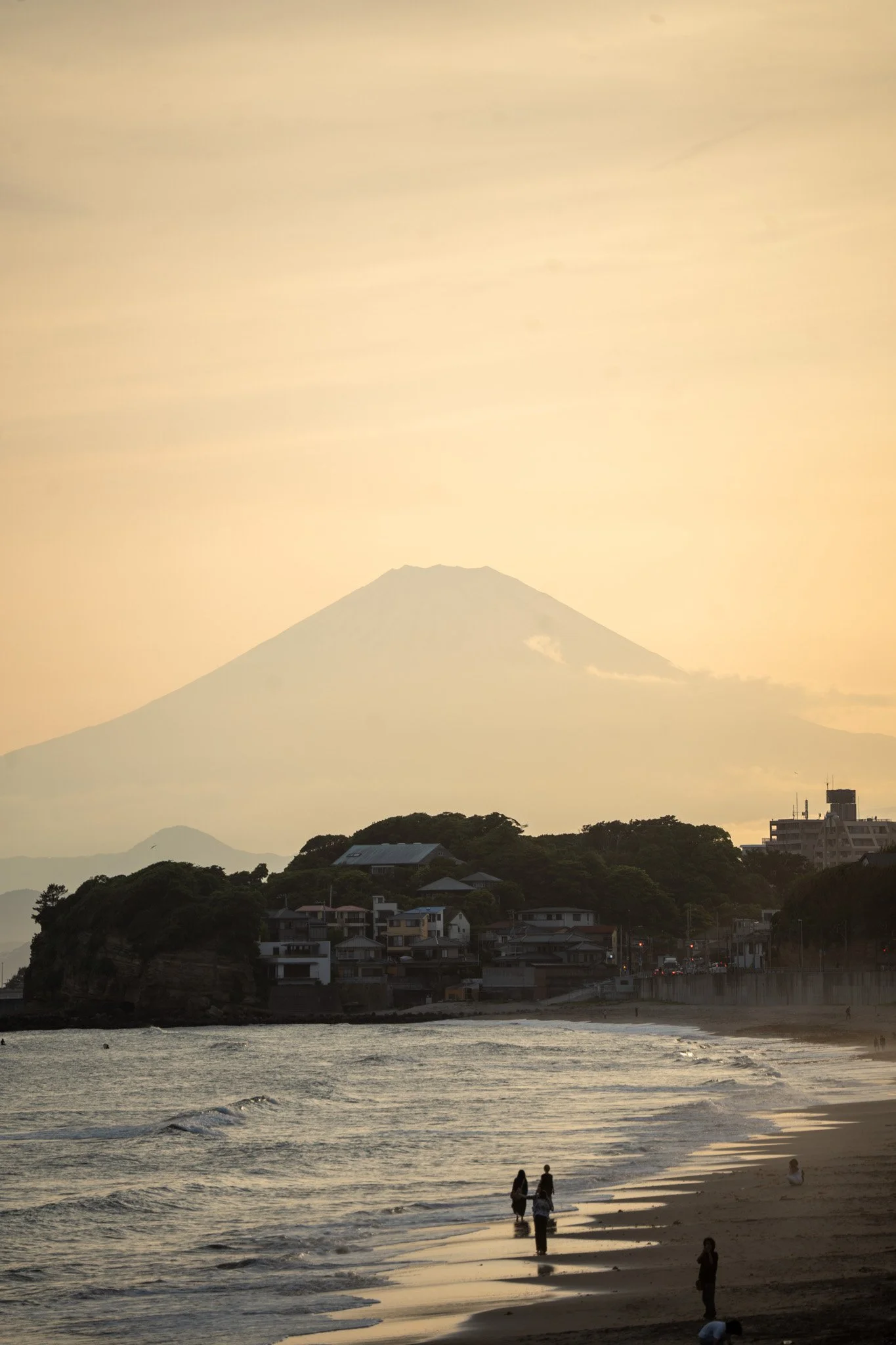 Beach scene at sunset with Mount Fuji in the background, and people walking along the shoreline.