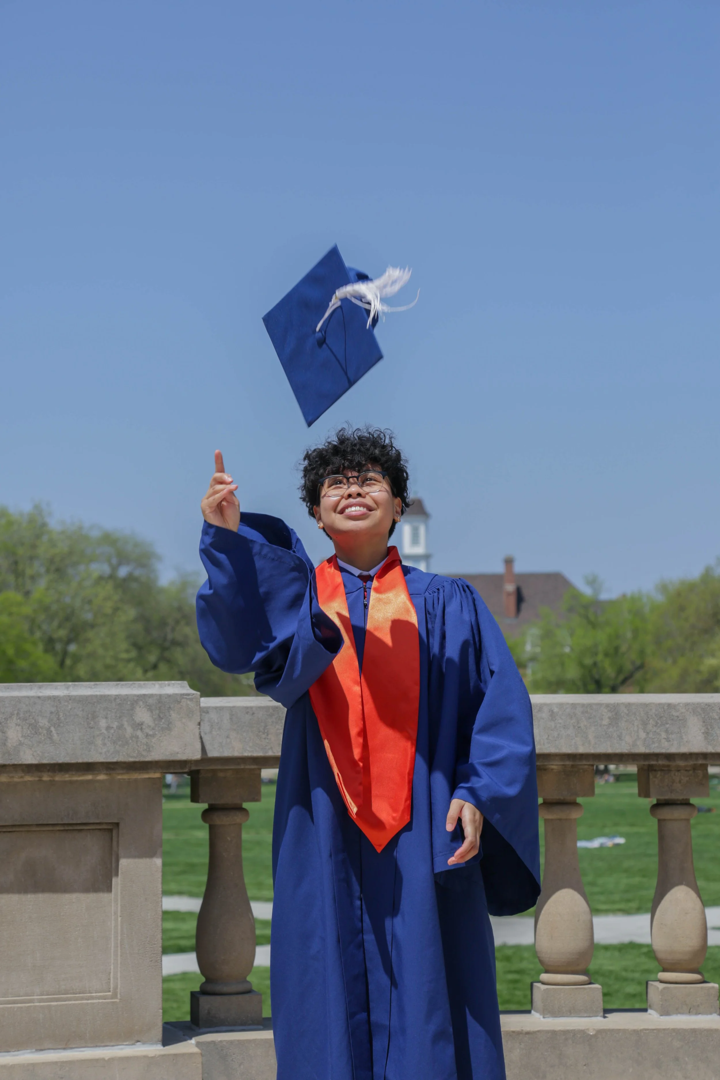 A young graduate in a blue gown and orange stole is outdoors on a bright day, tossing her graduation cap into the air with a joyful expression, standing by a stone railing with a school building and green trees in the background.