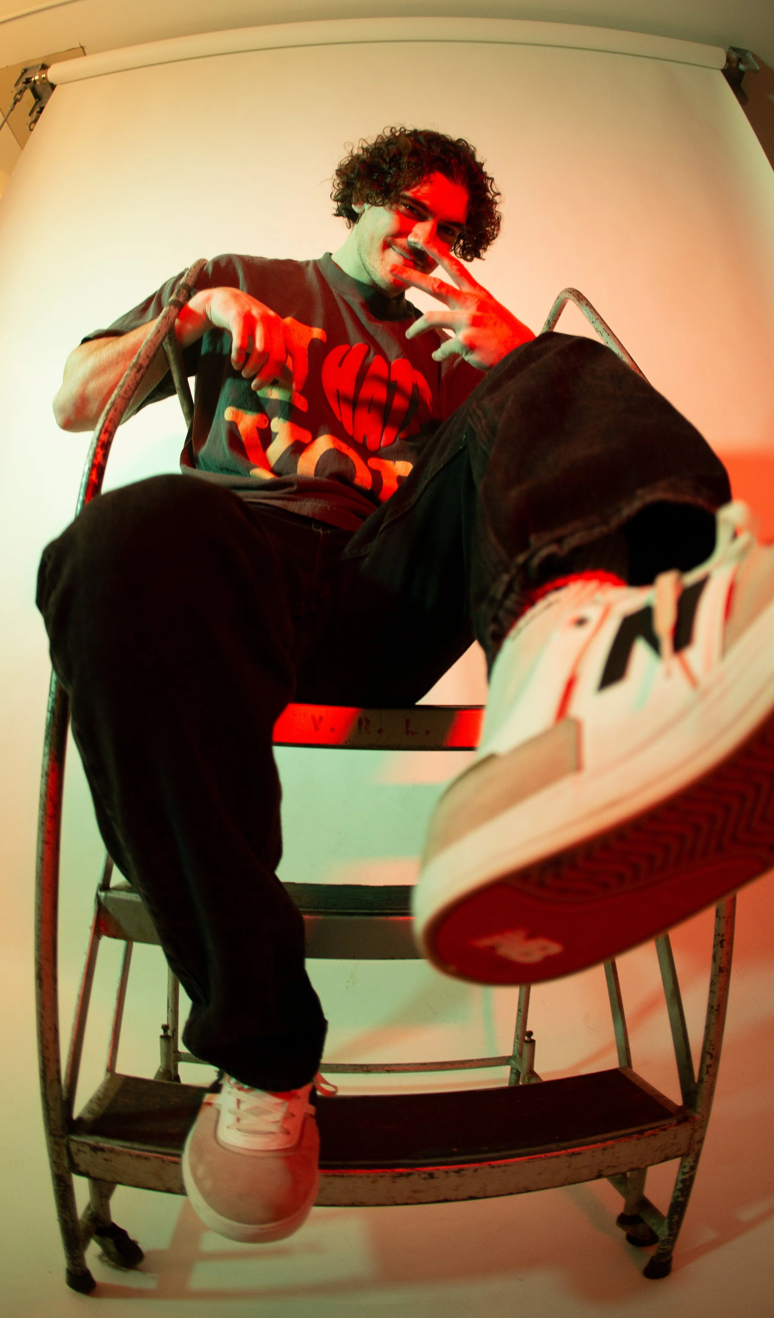A young man with curly hair smiling and making a peace sign with his fingers, sitting on a ladder against a plain background, wearing a dark t-shirt and sneakers, shot from a low angle.